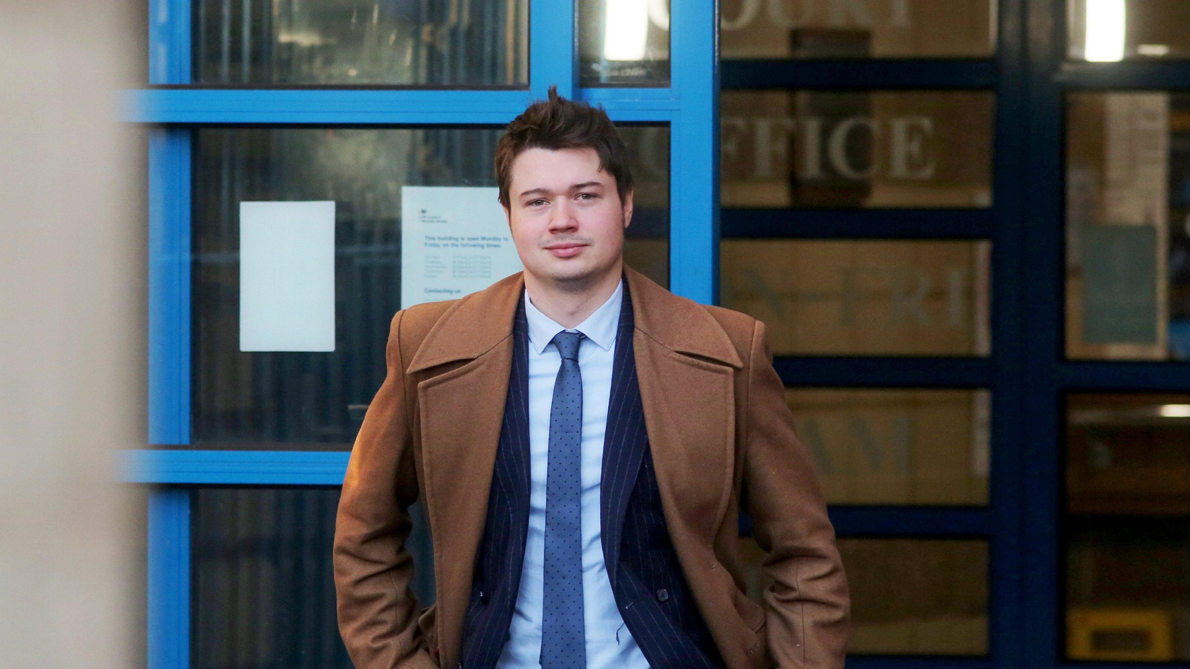 A man in a light brown overcoat with a navy suit jacket, blue tie and light blue shirt underneath. He has short brown hair. He is facing the camera with his hands in his pocket. Behind him is a blue door that is partially open