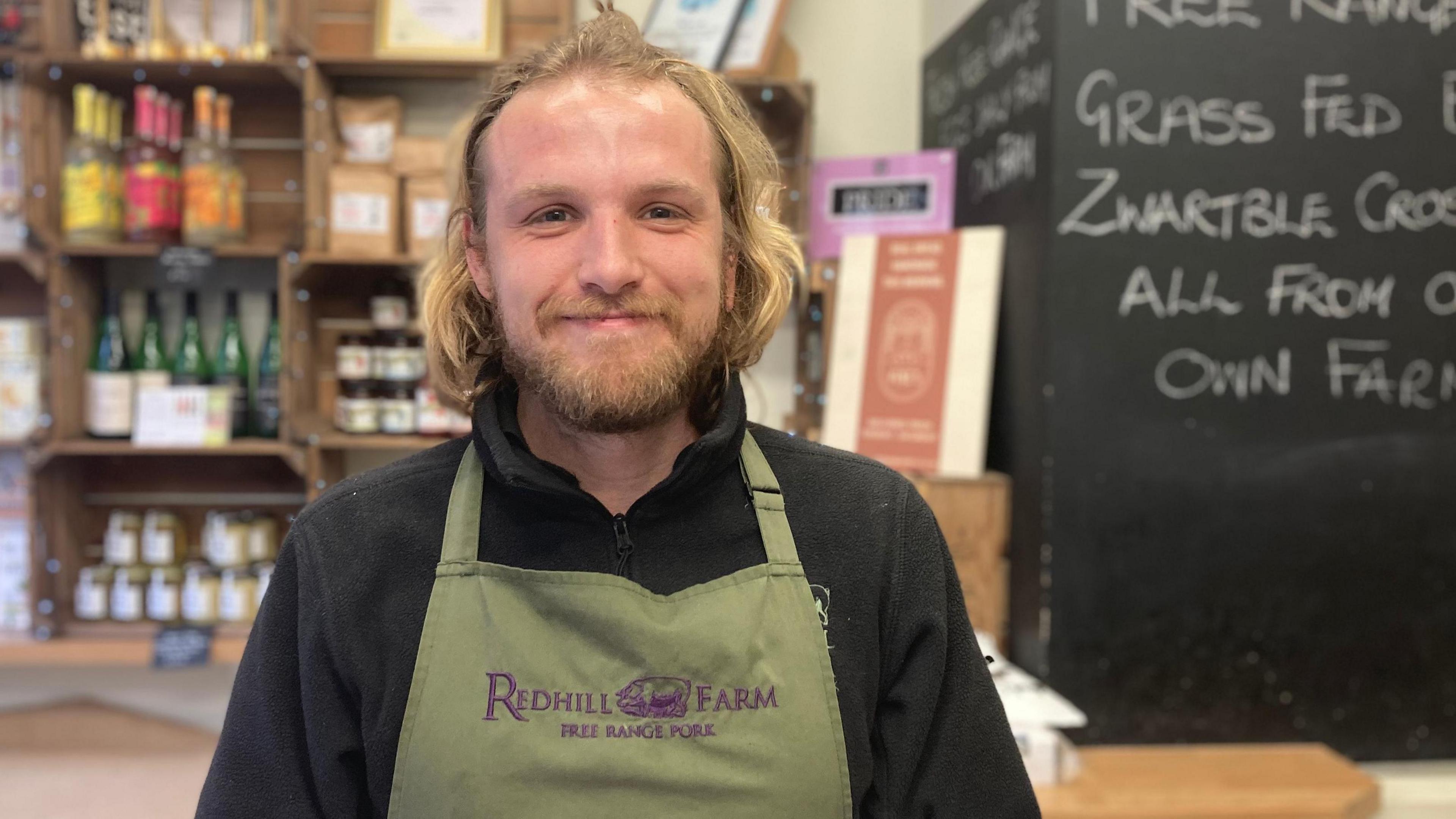 A smiling Ashley Delaney wearing a green tunic with "Redhill Farm Free Range Pork" printed on it. He is standing in front of a board promoting what is on offer in the shop