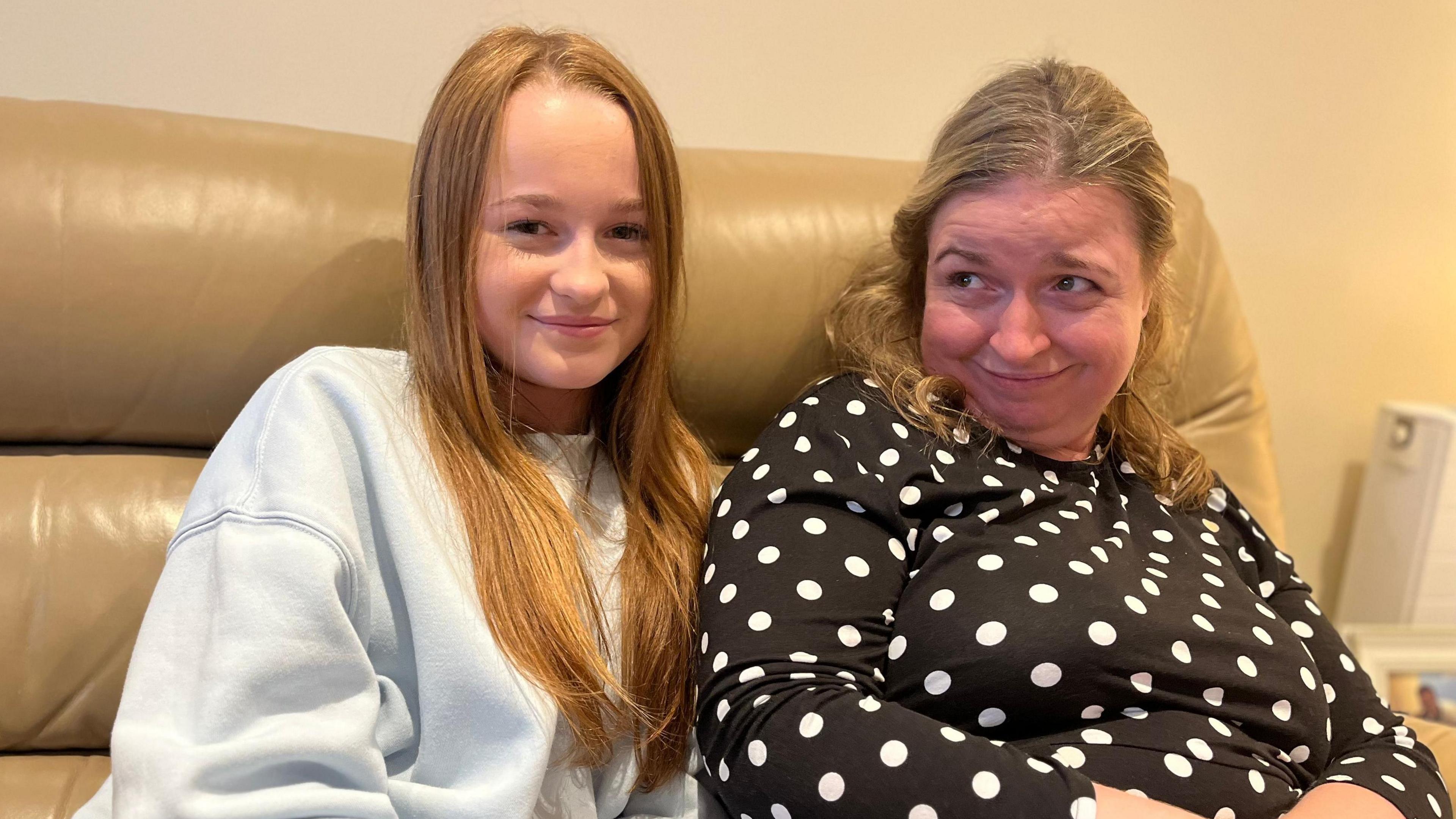 A teenage girl with long straight dark blonde hair, parted slightly off-centre, and wearing a light blue crew neck sweatshirt, sits on a tan coloured leather sofa. Next to her is a woman in her forties, who has dark blonde hair parted down the middle and shoulder length, slightly curled, worn in a half up, half down style. She is wearing a black, long-sleeved dress with white polka dots. The young girl is smiling into the camera, while the woman is also smiling but looking off to the left of the frame. The wall behind the sofa is painted a peach colour.