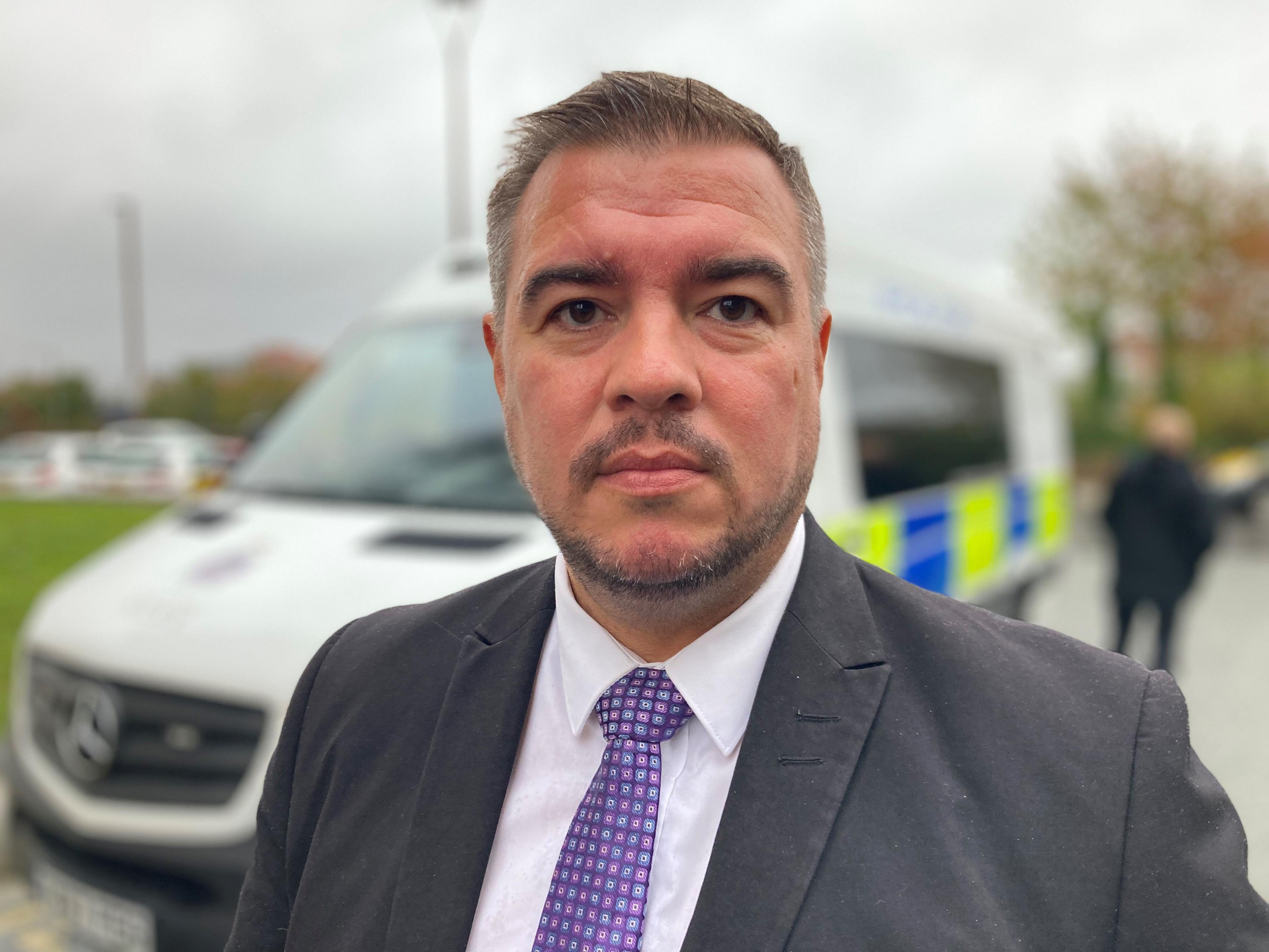 Det Ch Insp Stuart Hodgson is looking into the camera with a neutral expression. He has short brown hair and a dark beard. He is wearing a grey suit with a purple tie. A police van can be seen in the background.