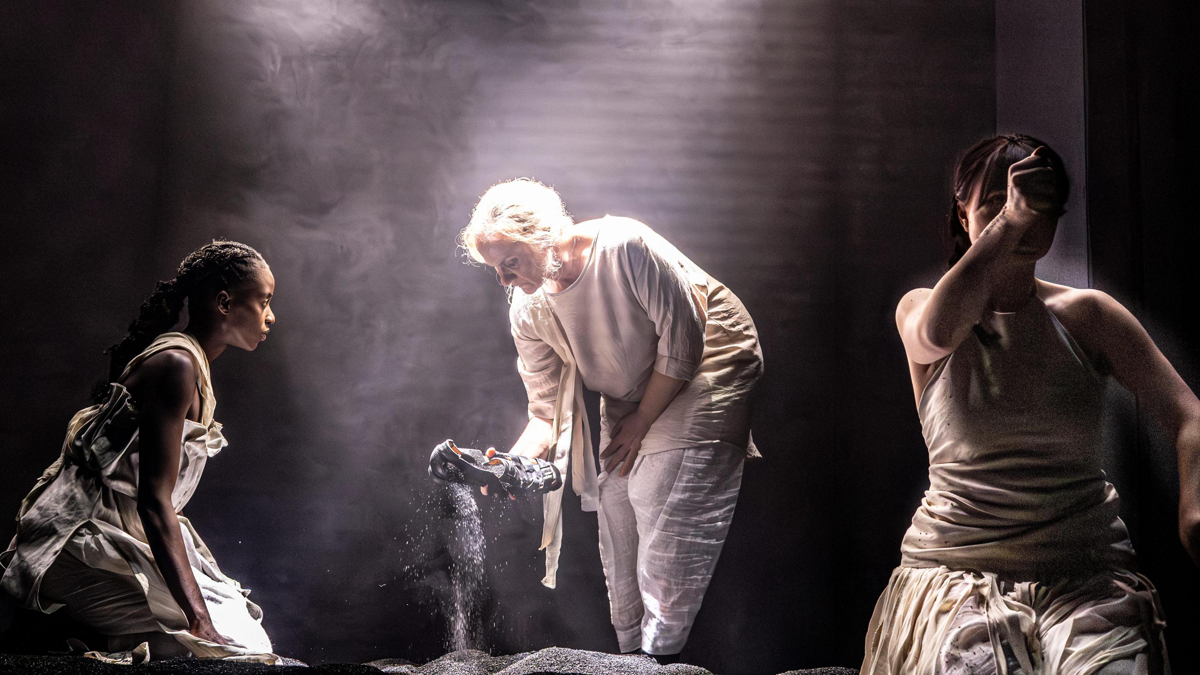 A scene in a play with three actors dressed in minimalist, loose clothing. A woman in the left is crouched watching another woman pulling a sandal from a piles of black dust. To her right another woman is holding an outstretched fist to the camera.