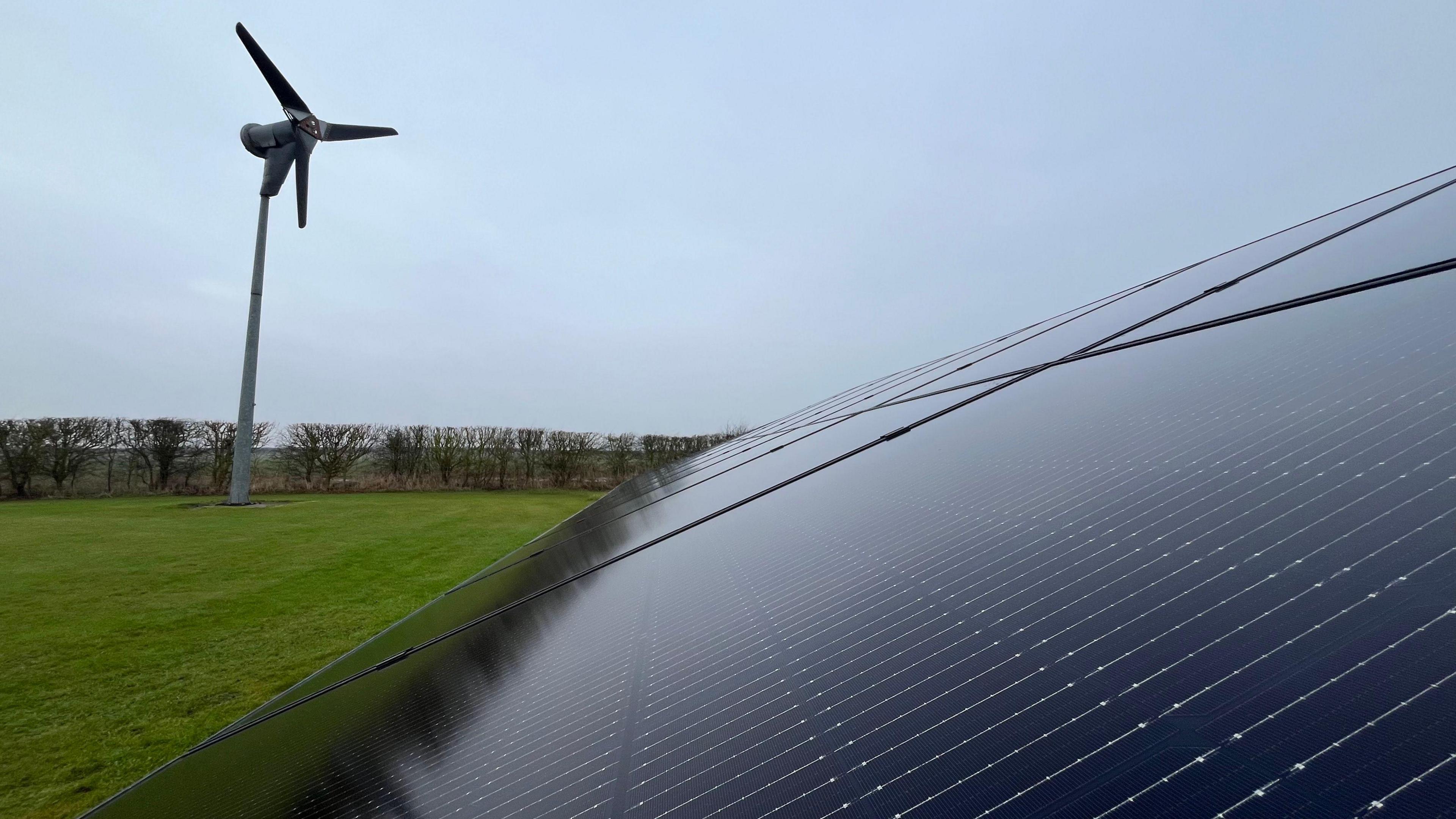 A ground-mounted solar array and wind turbine in a grass field.