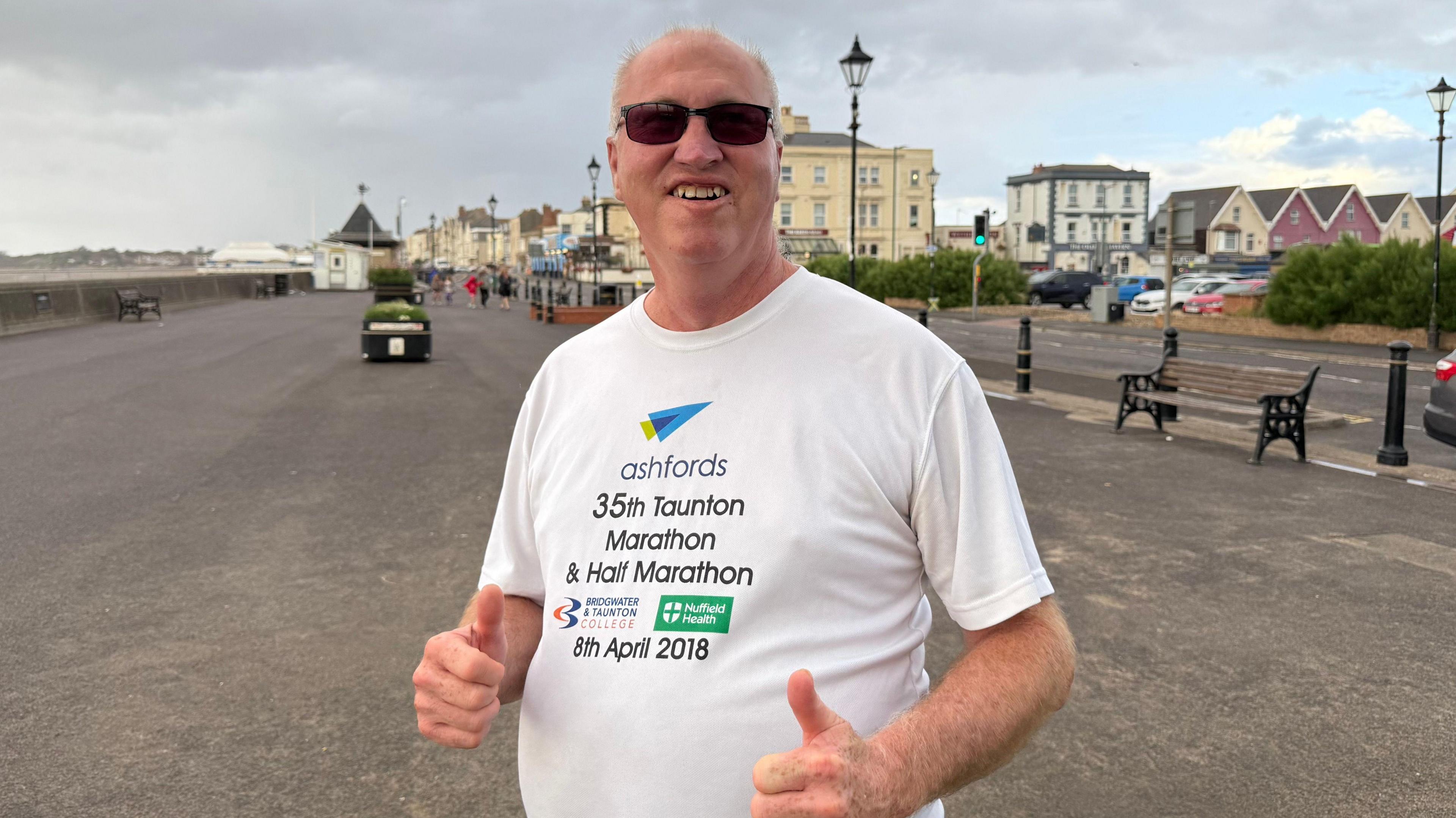 Mr Vickers in a Taunton marathon t shirt as he trains in Burnham. He has sunglasses on and has both thumbs up as he smiles at the camera. In the background, you can see the beach road in Burnham.