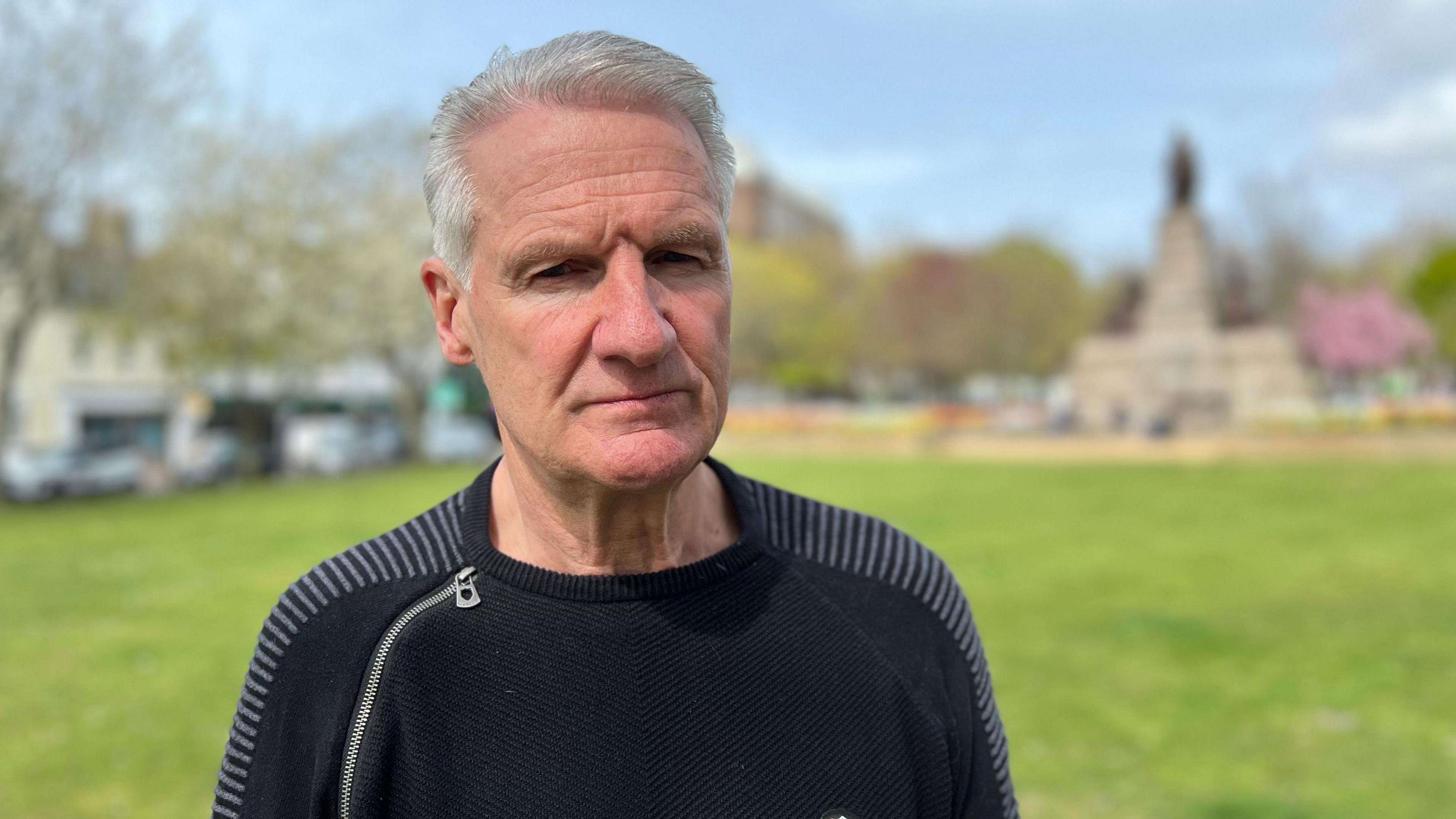 Tom Binet looks very serious as he looks towards the camera. he is wearing a black top with a zip on his right shoulder. He is standing in a park with a statue in the background. 