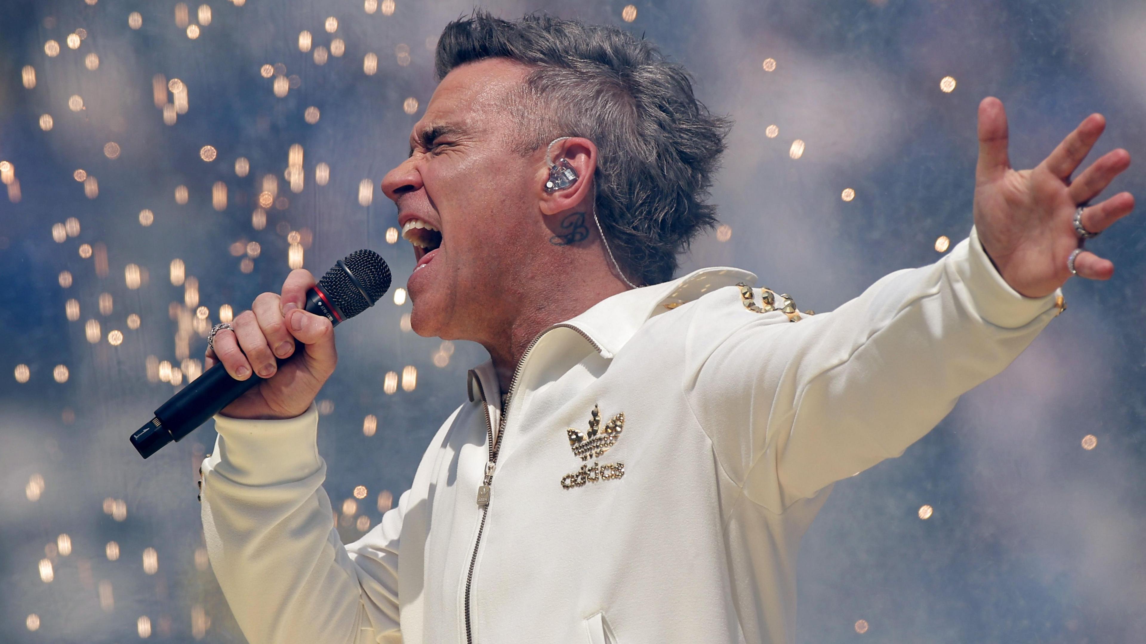 Robbie Williams performs in a white Adidas tracksuit with gold trim. He is holding a microphone and singing emphatically as pyrotechnics fall behind him.
