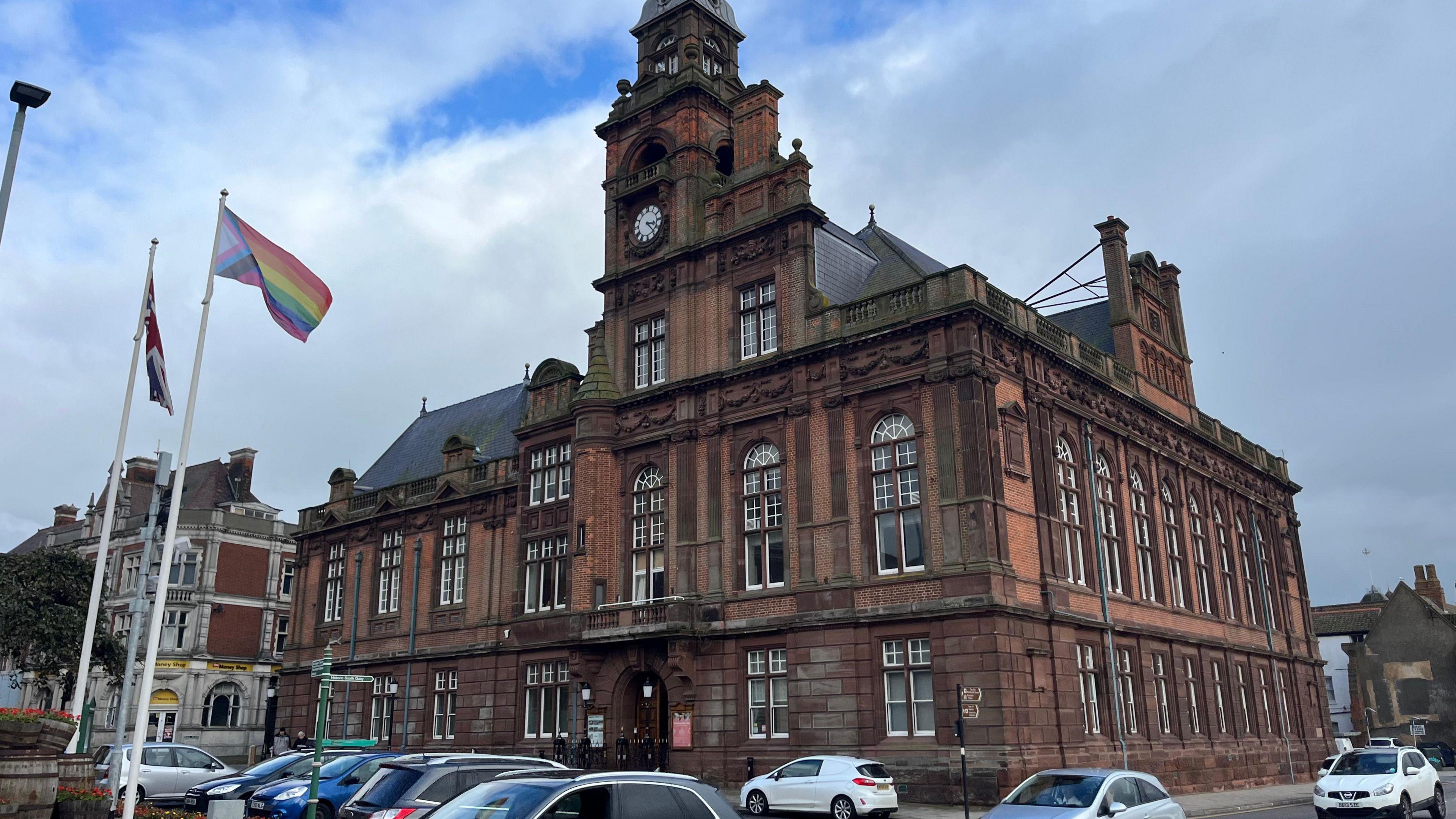 A large brick building with a clock tower. Cars are parked in front of it and two flags- a rainbow-coloured and a red, white and blue union flag are flown from two poles.