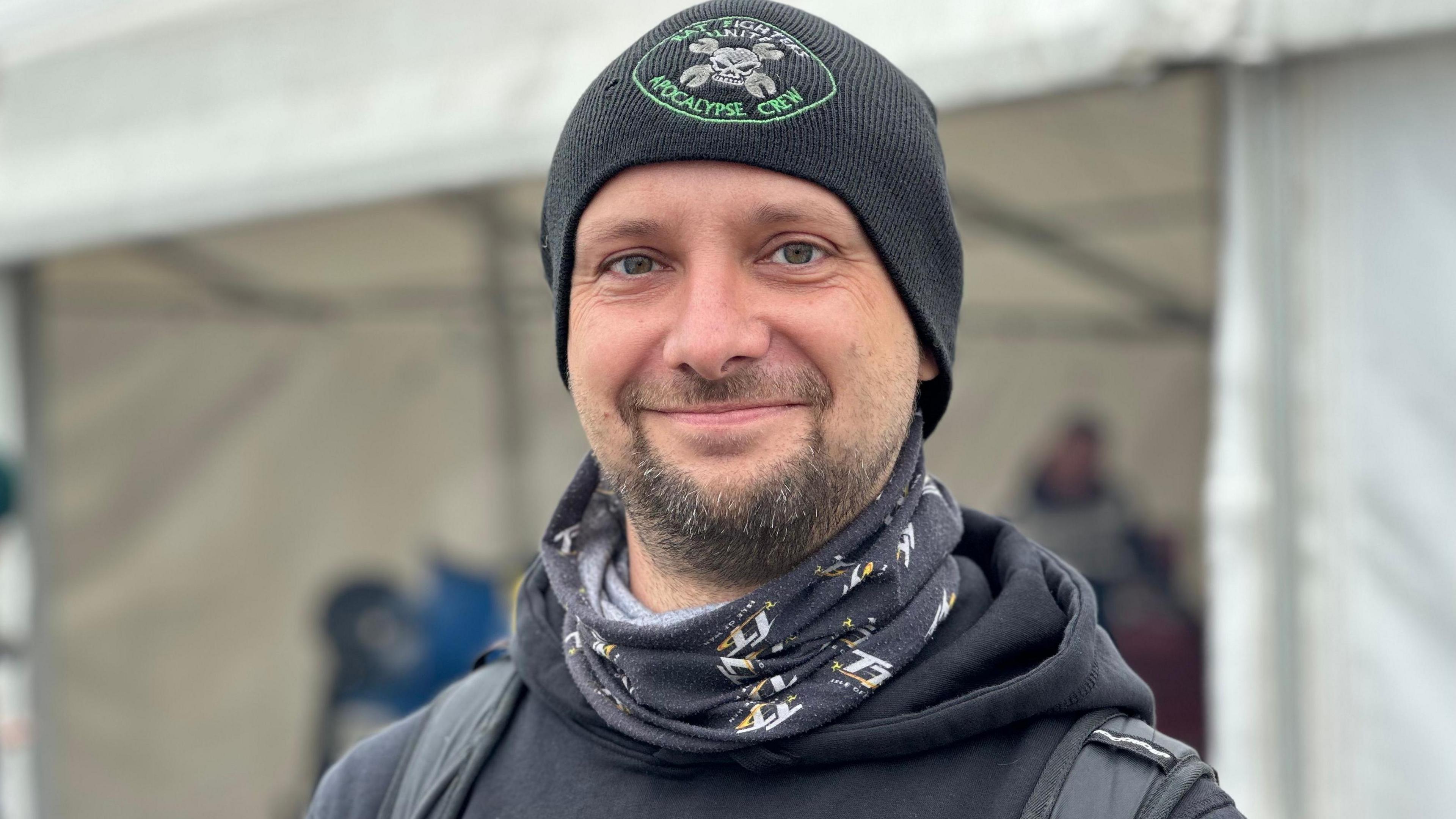 Stefan Hahn wears a black woollen hat and black hooded jumper, he smiles in front of a white marquee which is blurred out in the background.