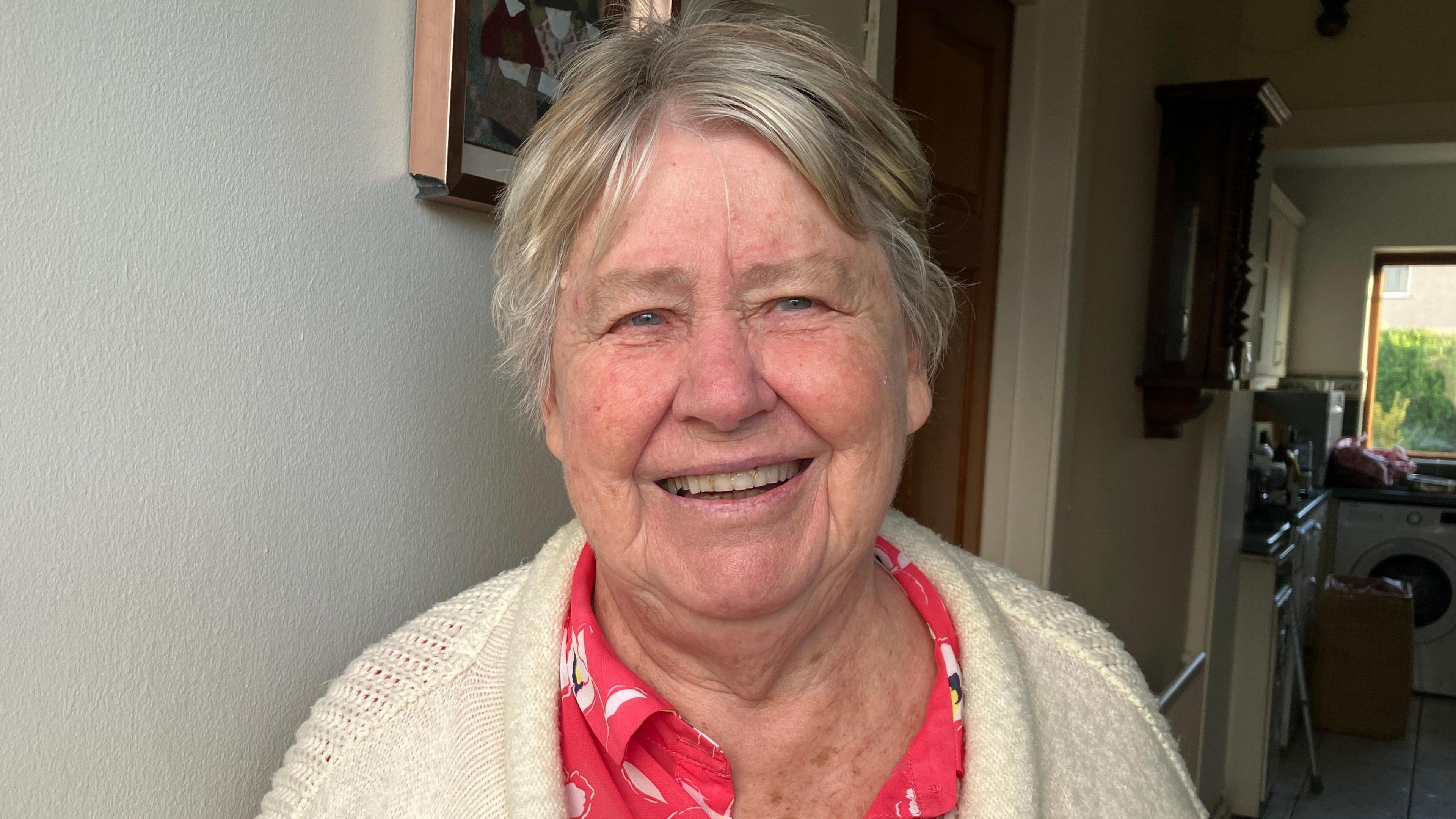 Menyw yn gweni at y camera ar ei stepen drws. Mae ganddi wallt gwyn ac yn gwisgo ffroc a cardigan gwyn. Yn y cefndir mae'n bosib gweld ei chyntedd a'i chegin. 