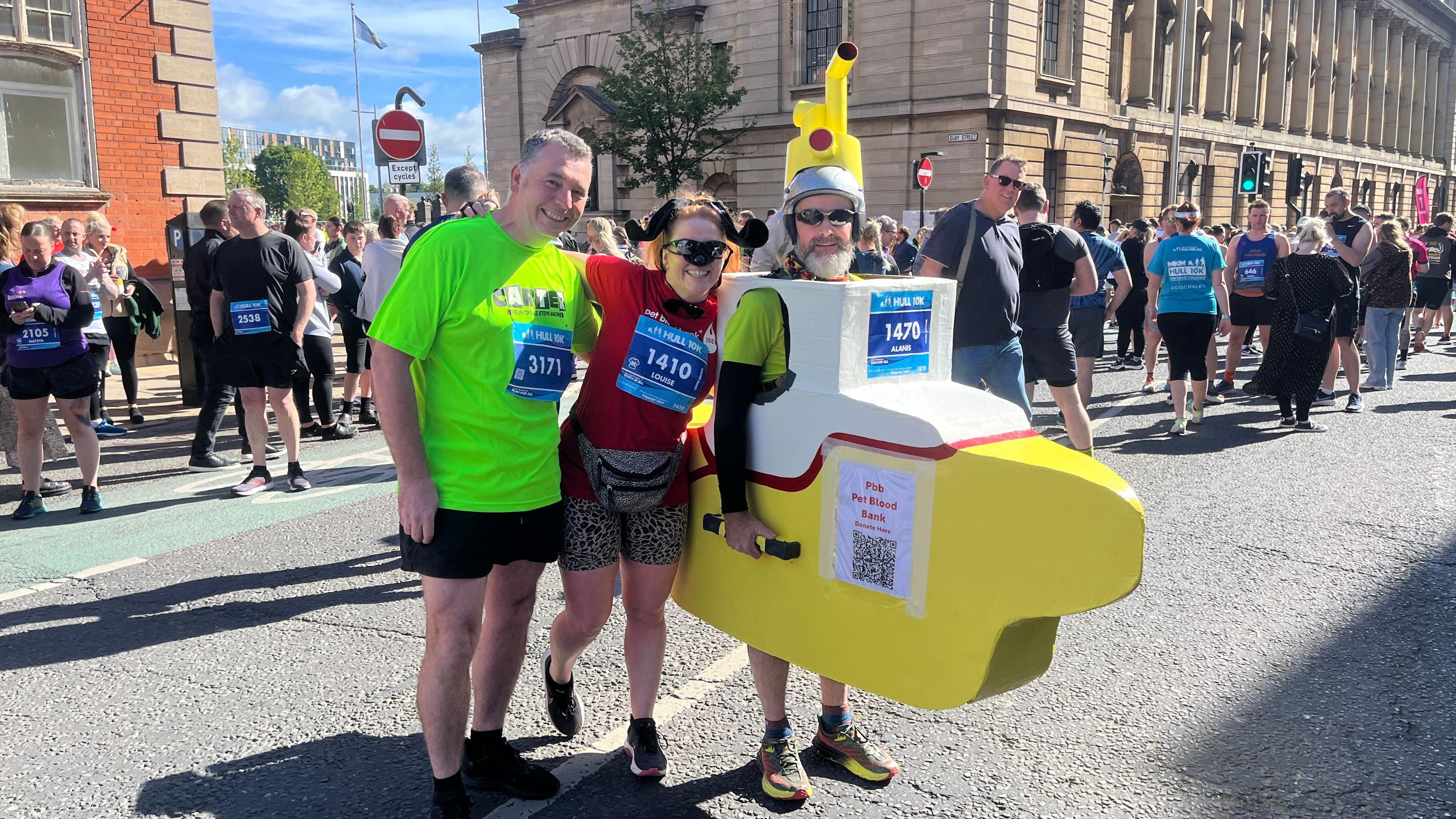 Three runners, two men and a woman, pose at the start of the Hull 10k run. The man on the left is wearing a fluorescent yellow top and black shorts. The woman in the centre is dressed as a dog and is wearing black sunglasses and leopard print shorts. The man on the right is dressed as a yellow submarine. Other runners and the Hull Guildhall are visible in the background.