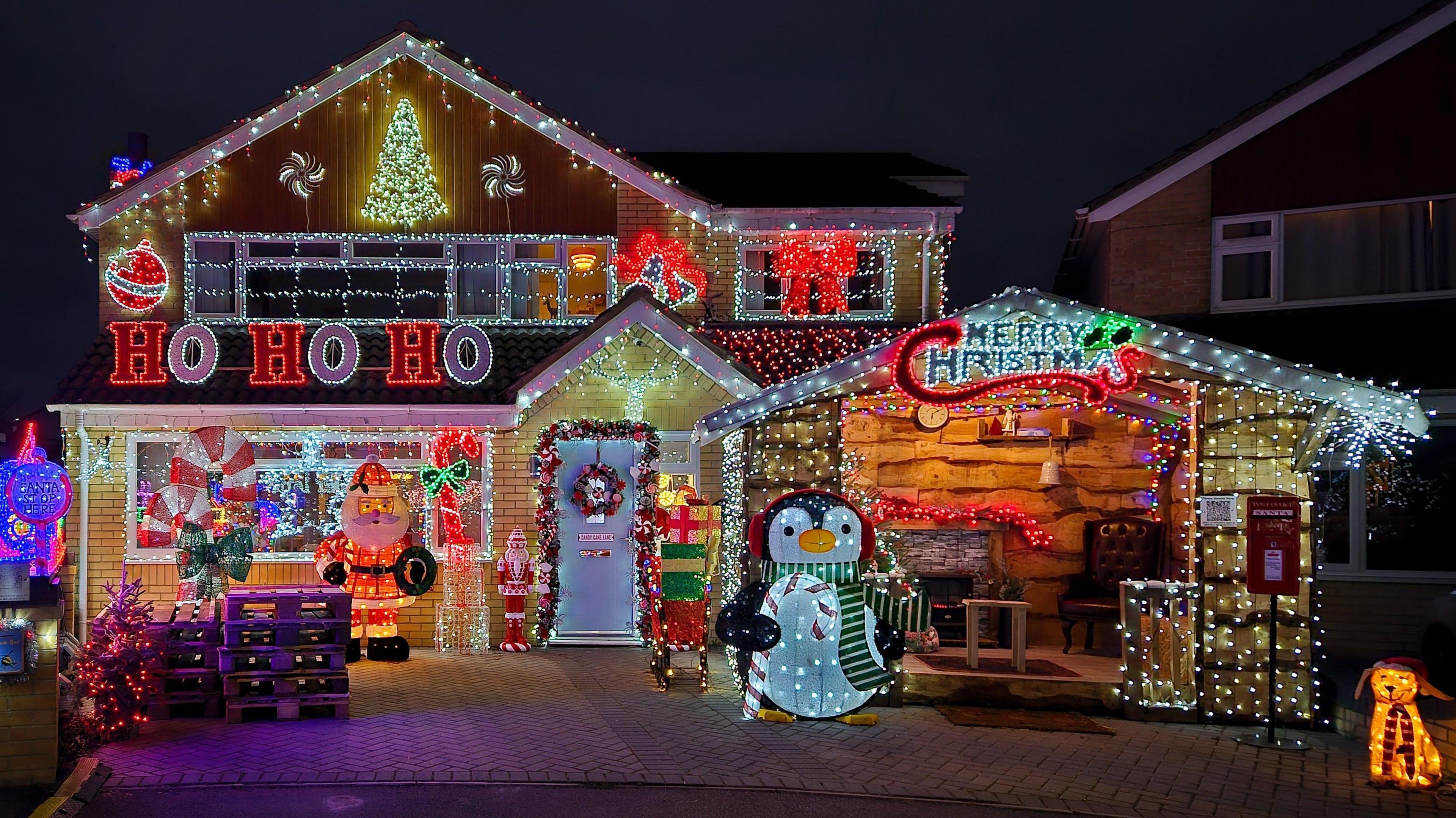 Sarum Avenue in Melksham, Wiltshire. A house is covered in Christmas lights. There are large figures including a Santa, a penguin, presents and a dog which are also lit up. 'Ho ho ho' is spelled out in lights, as is 'Merry Christmas' 