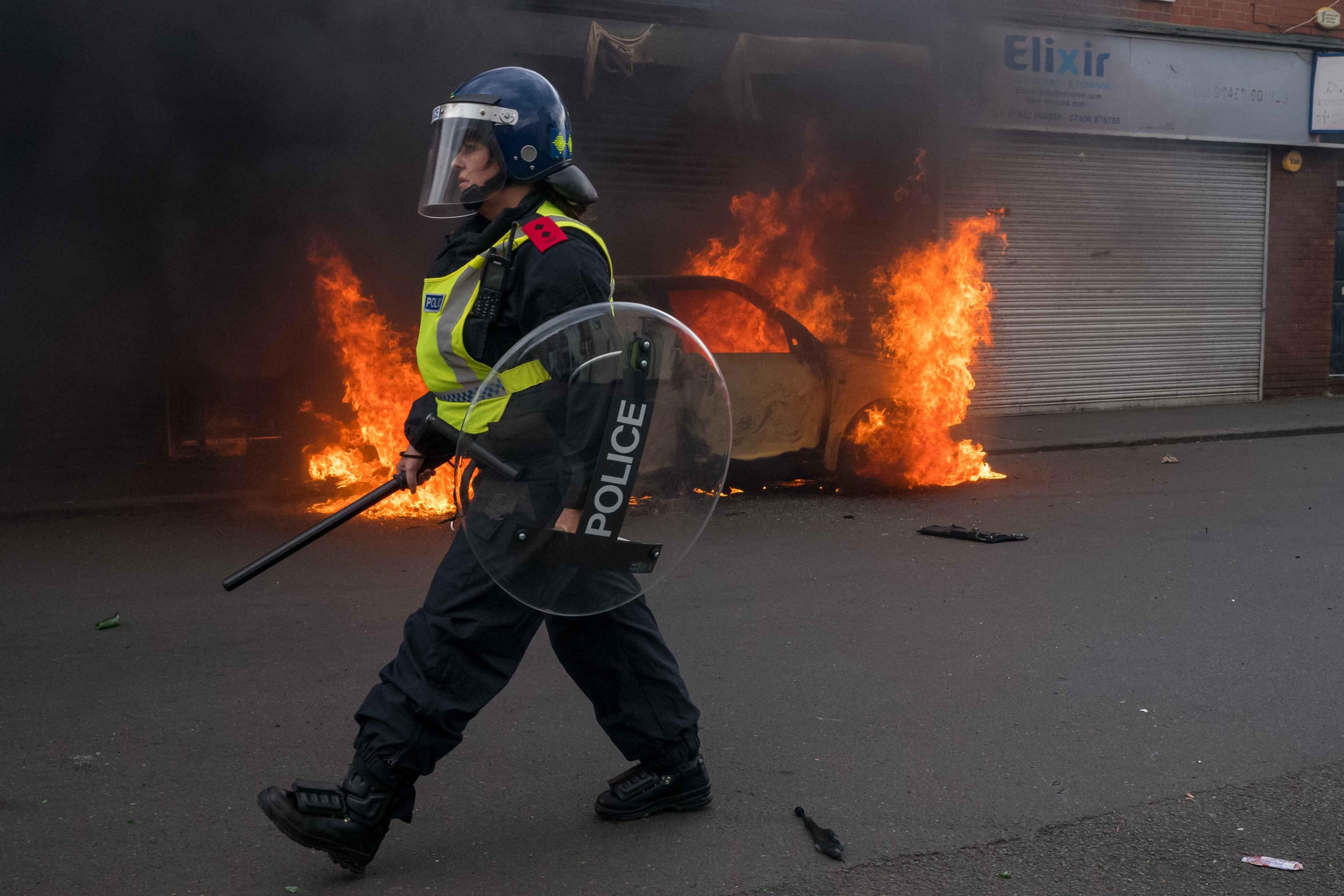 A car burns on a road during a riot. A police women , wearing riot clothing and a shield is standing in front of it. 