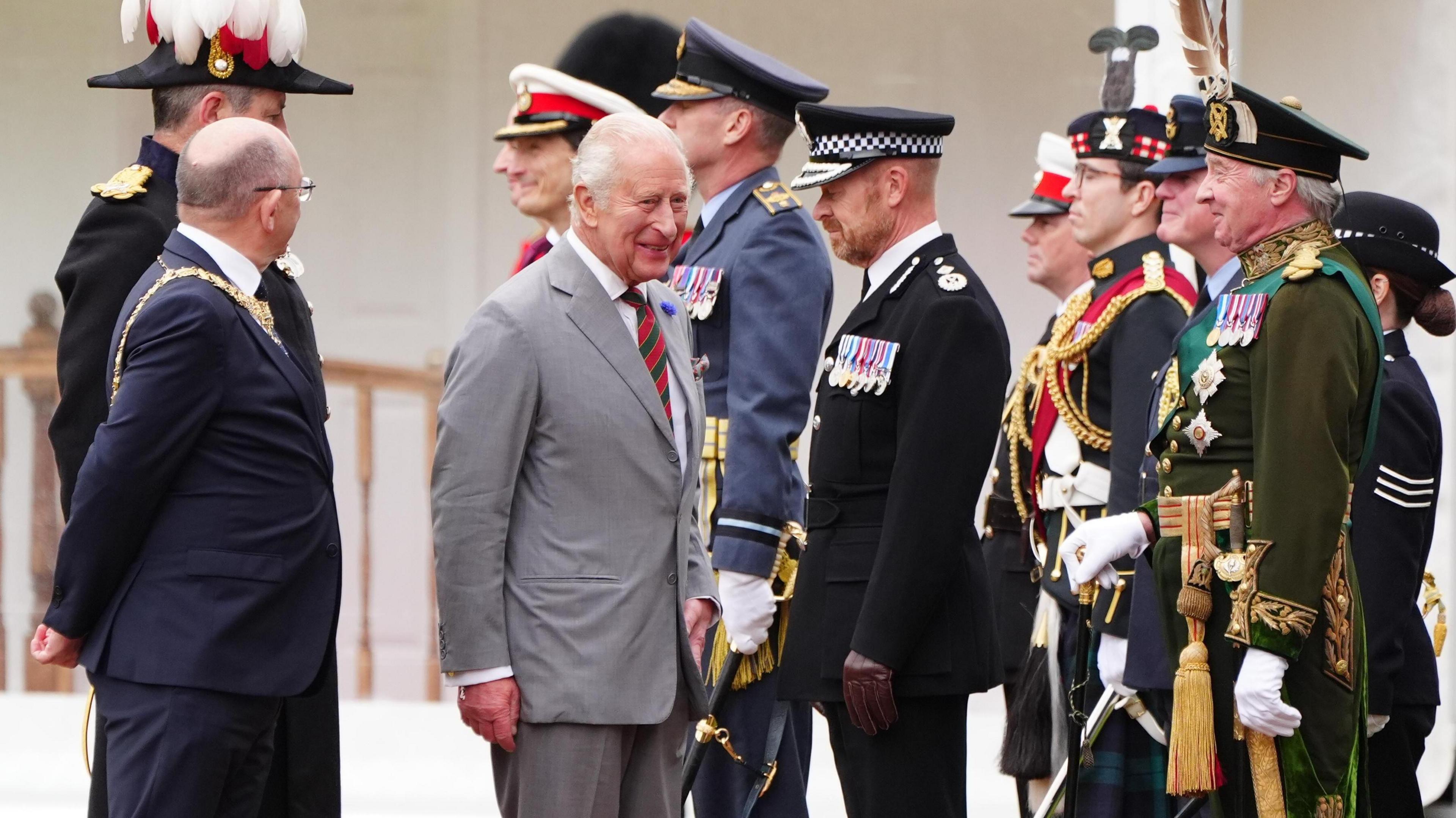 King Charles III wearing a grey suit and white shirt walks with the Lord Provost of Edinburgh Robert Aldridge, who wears glasses and a dark-coloured suit. The pair are greeted by uniformed figures.