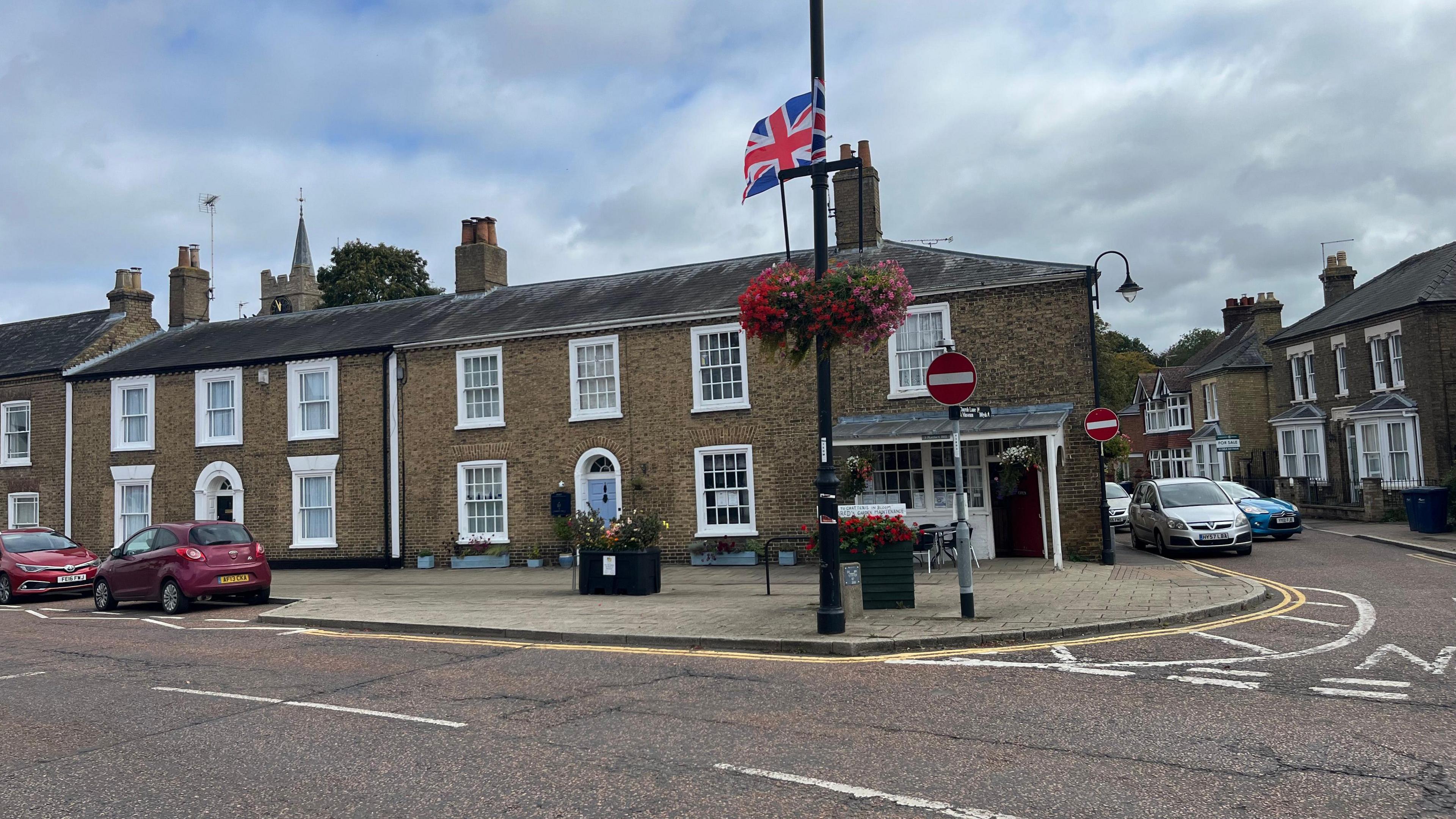 A terrace of Victorian buildings in a town centre, with some cars parked along the streets. There is a union flag placed above a couple of colourful hanging baskets stuffed with flowers.