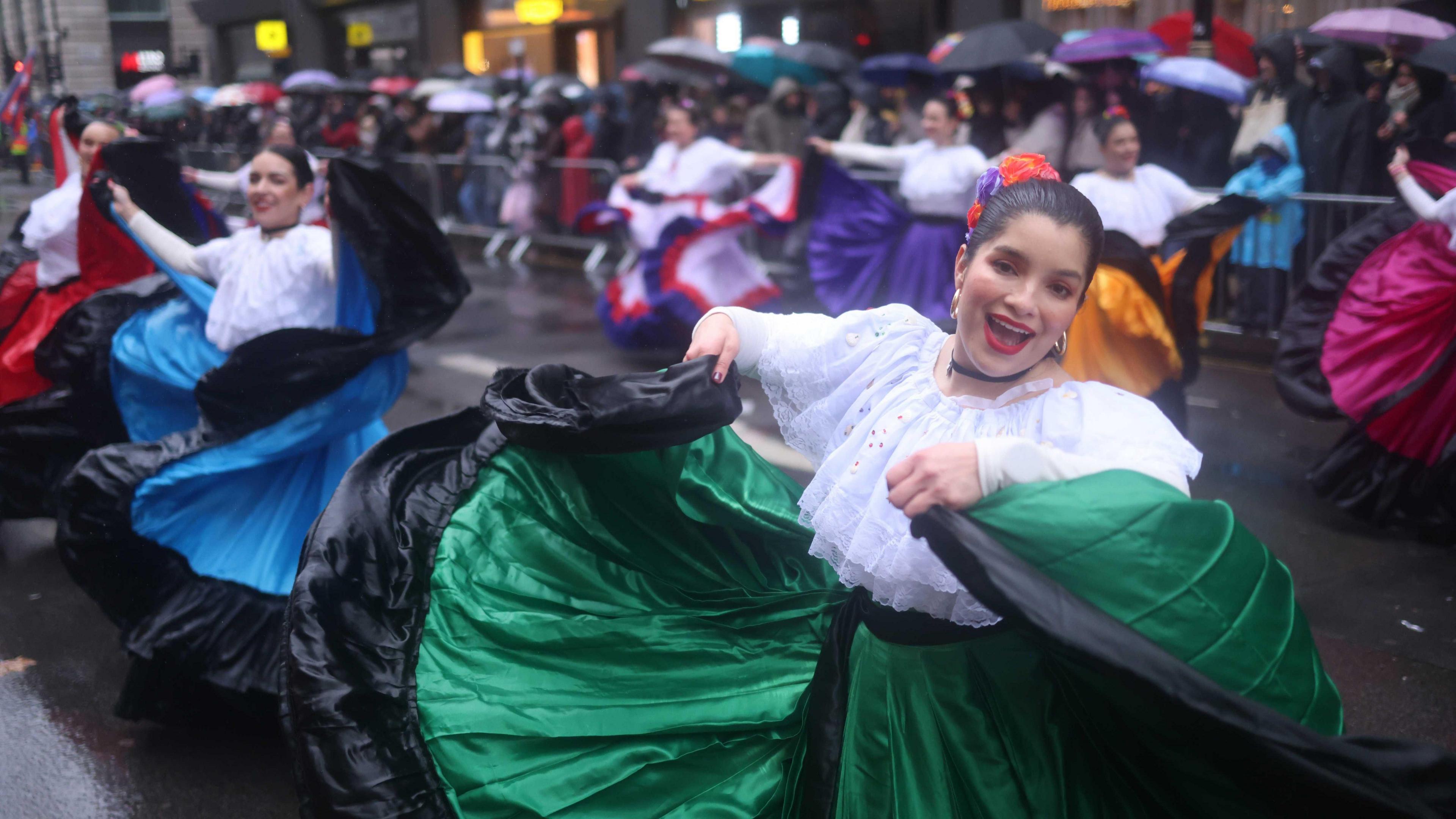 A woman in traditional Spanish attire, a flowing green and black skirt and white blouse, smiles as she dances and looks into the camera. 