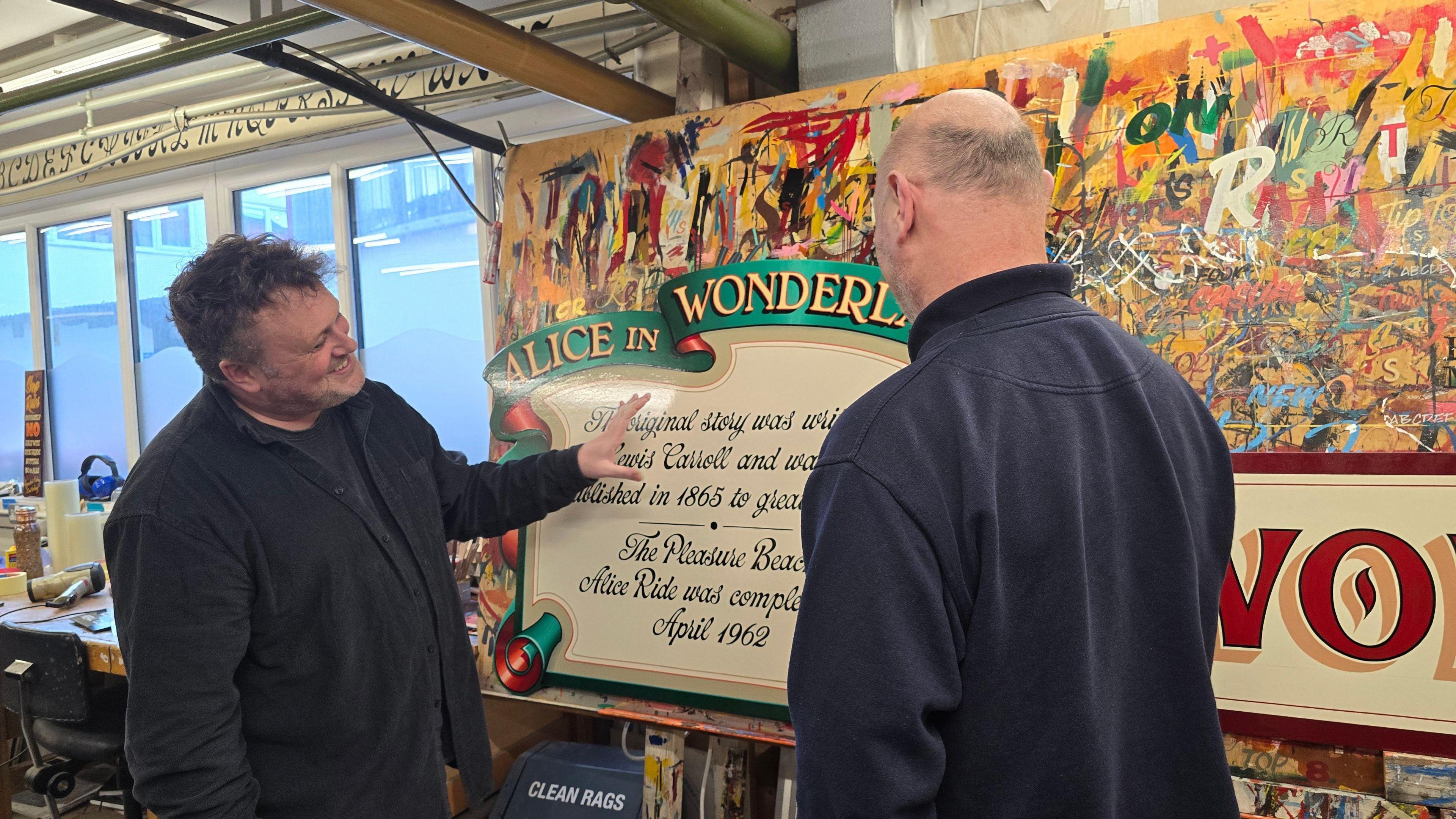 A man smiles as he stands and appreciates a colourful Alice in Wonderland fairground sign as it stands in a workshop, a man on the right looks on at him and the sign 