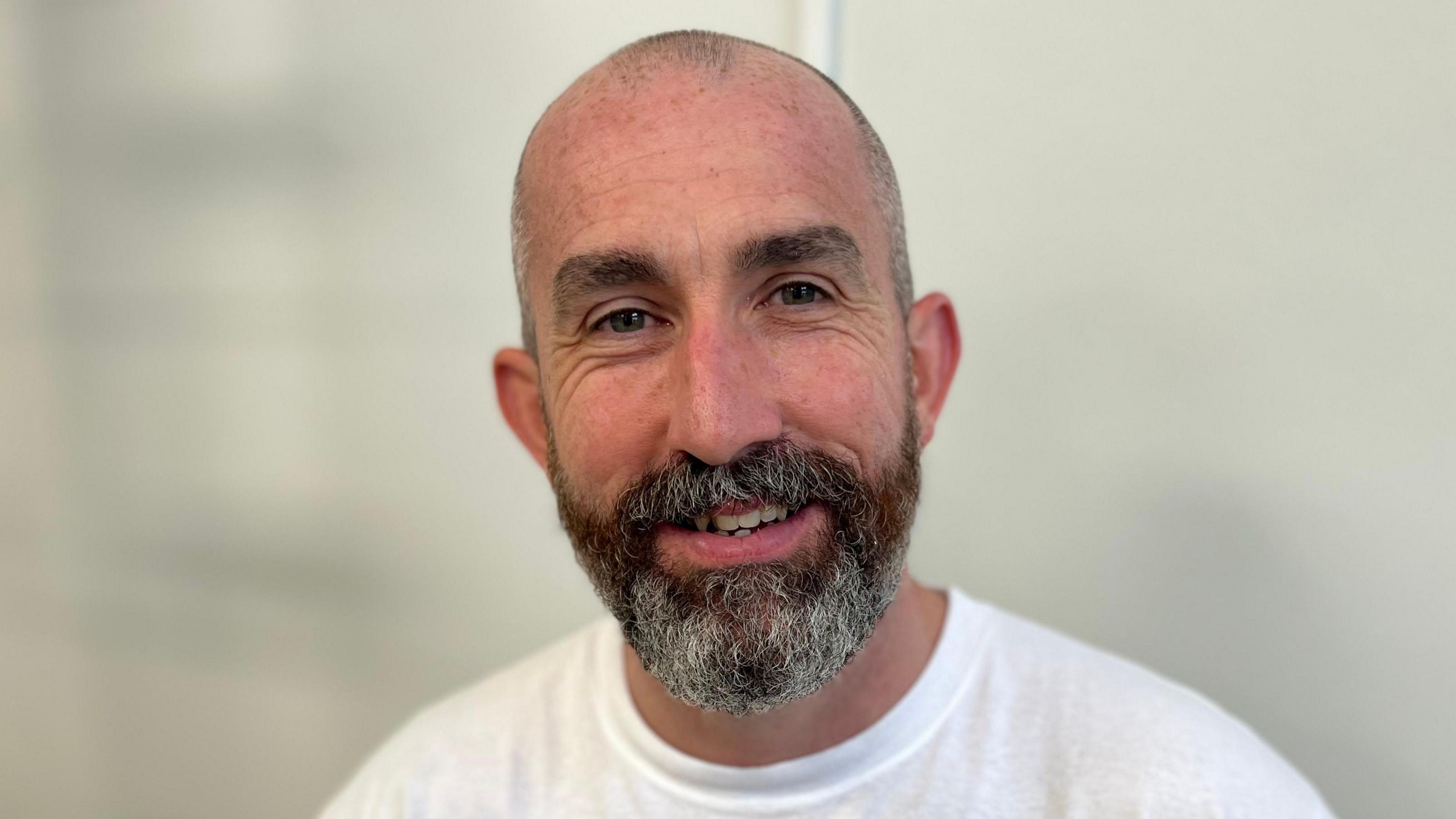 Gerard Donaghy is standing in front of a white wall, wearing a white t-shirt and is smiling at the camera. He has a bald head and a dark beard with grey hair through it.