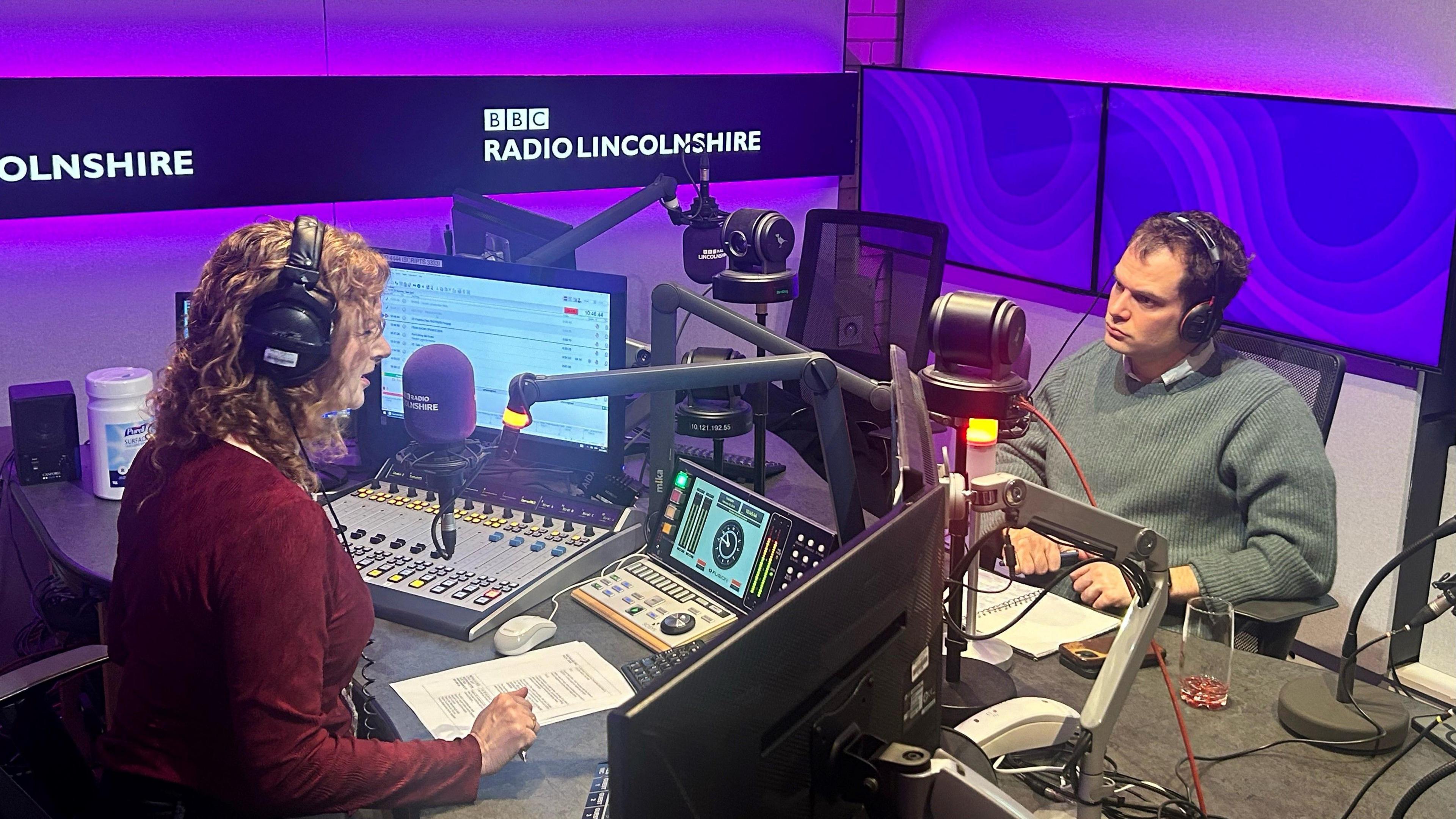 A BBC radio studio is lit with purple lights and has Radio Lincolnshire written on boards on the walls. The presenter is a woman with curly ginger hair wearing headphones and a red top, sitting in front of a microphone. Sat opposite her is a man with short brown hair wearing a blue jumper who is also wearing headphones.