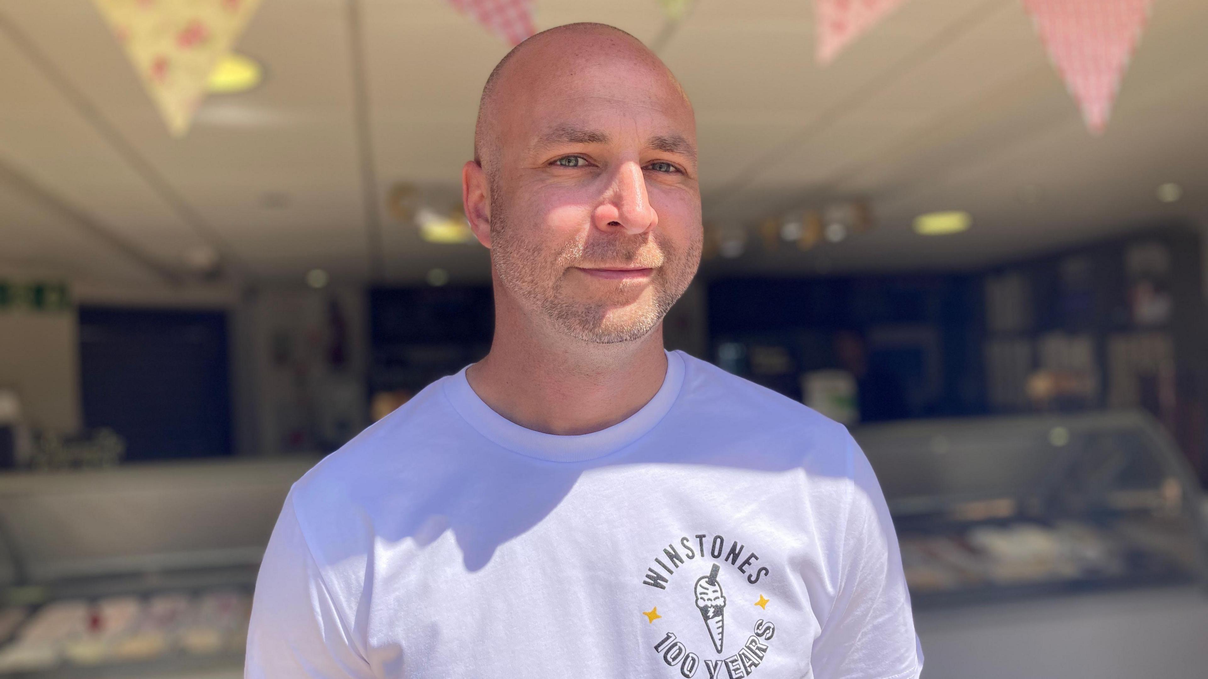 Tom Veal is wearing a white T-shirt with the words "Winstones, 100 years" on it, and smiles at the camera. The ice cream counter can be seen behind him.