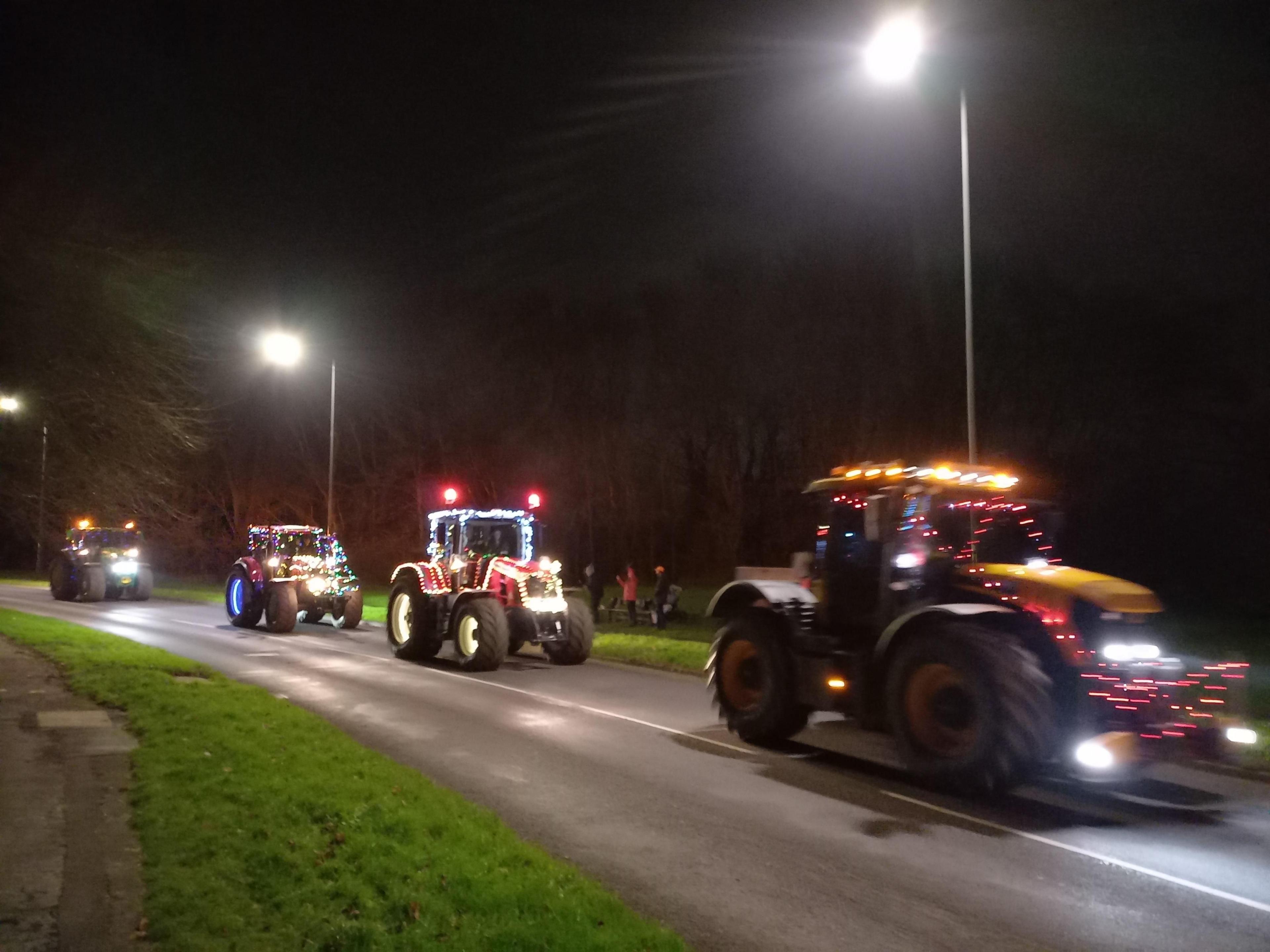 Four tractors driving along a road at night, all festooned in Christmas lights.