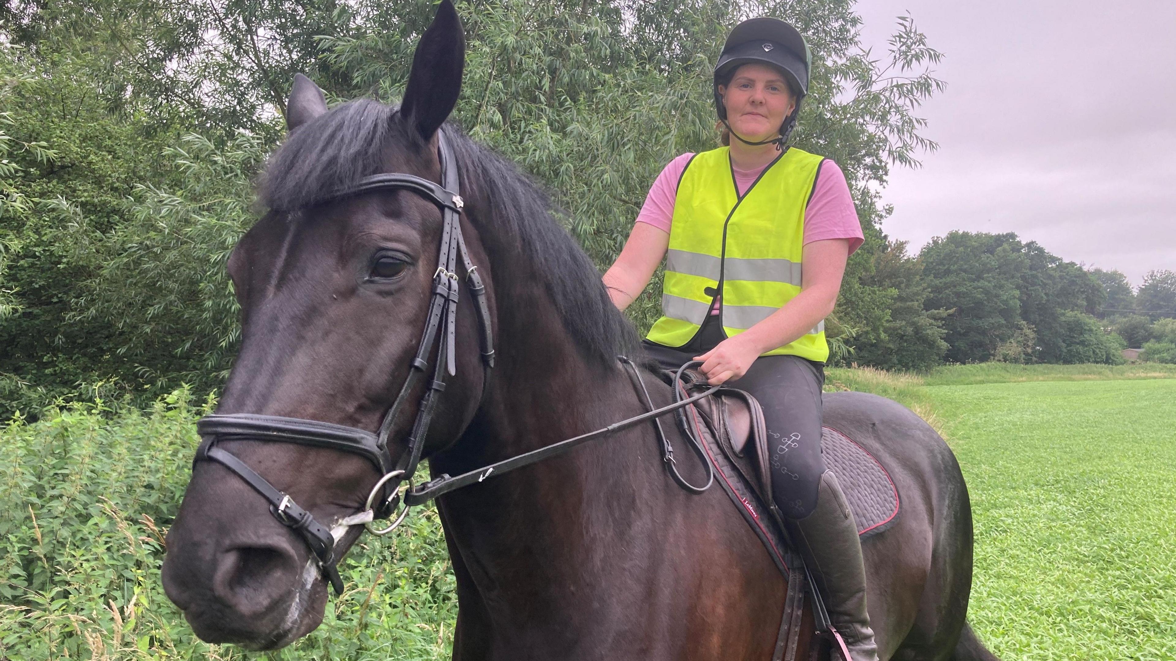 A female horse rider, wearing a pink t-shirt, black jodhpurs, a black riding hat and yellow hi-viz, is sitting on a dark bay horse in a field alongside a row of trees.