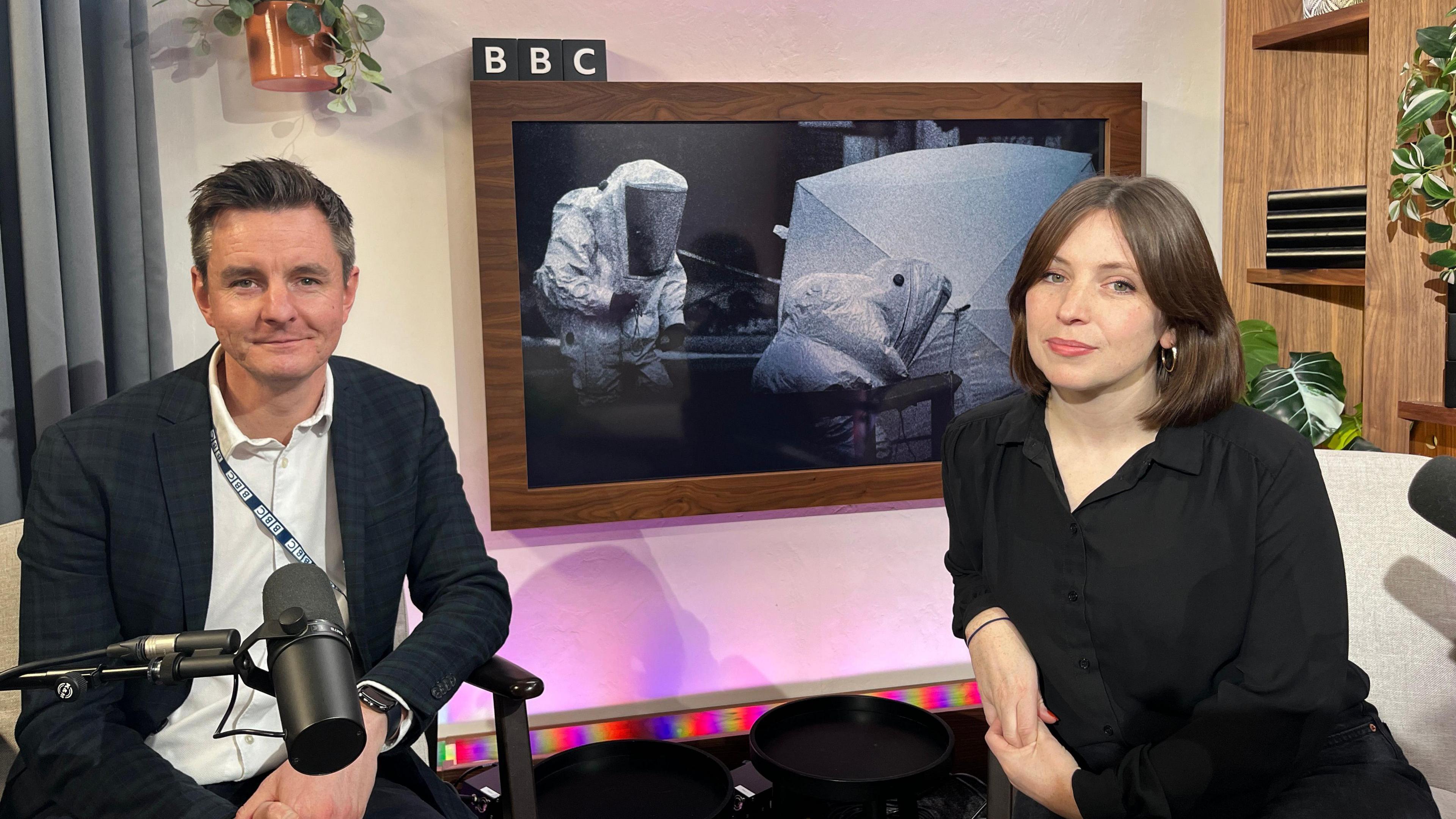 Andy Howard and Marie Lennon sat in their podcast recording studio. Andy has dark brown hair and is wearing a white shirt and blue checked blazer. Marie has a mid brown bob haircut and is wearing a black dress. 