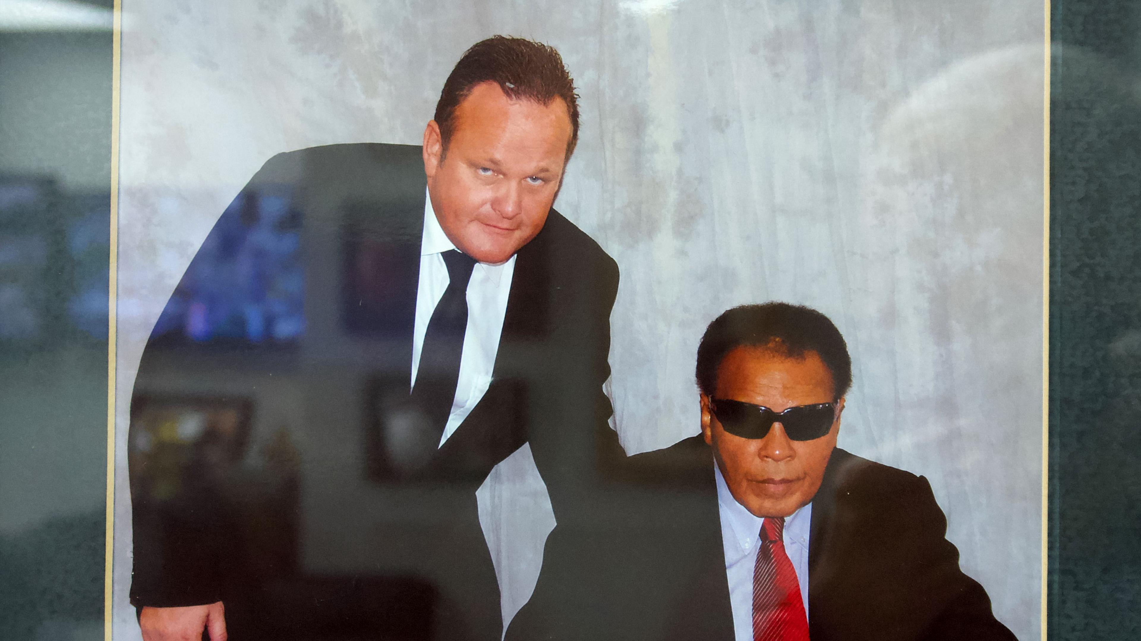 Two men in suits. One has a white shirt and black tie and is standing over a seated man wearing dark glasses and a red tie.