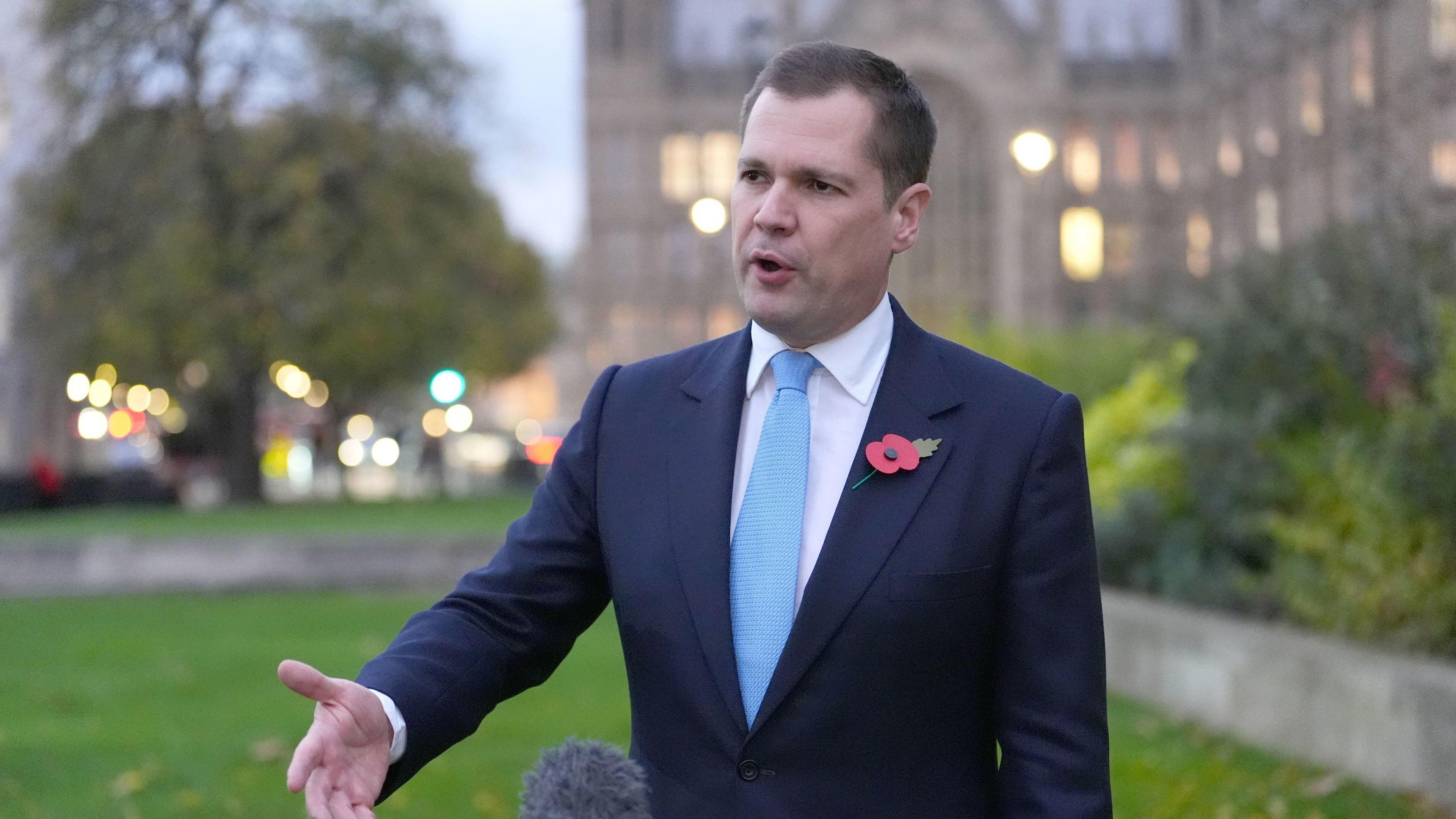 Robert Jenrick outside Westminster. He is wearing a navy suit and light blue tie. He has a poppy on his left lapel of his suit jacket. He has short brown hair and is clean shaven.