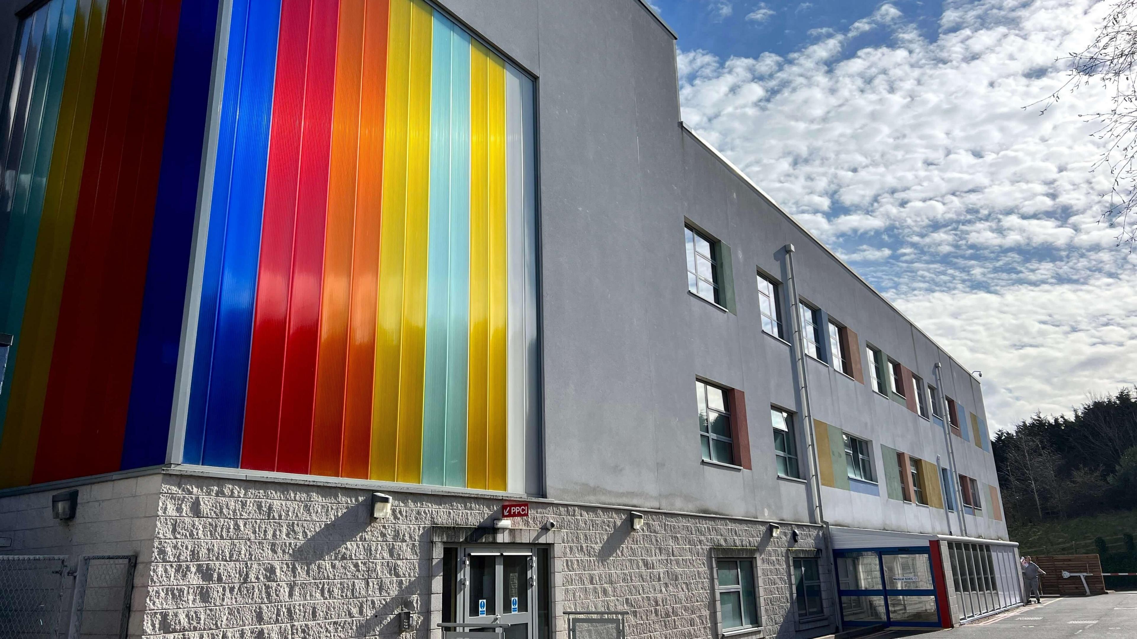 The outside of the Skylark children's ward at Kettering General Hospital which shows a grey, three storey bulding, which is cladded with a striped rainbow coloured decoration.