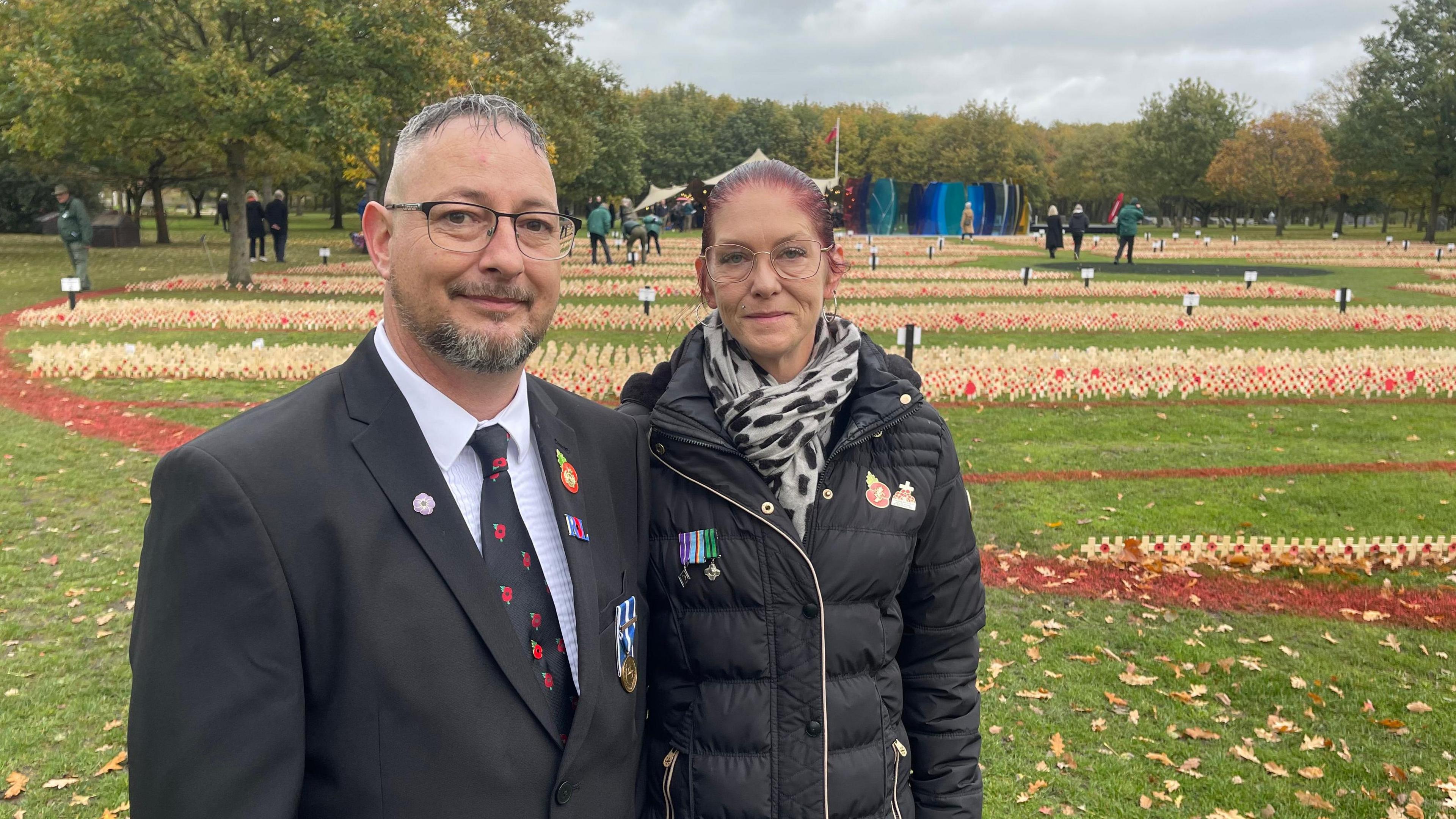 A man wearing a suit with a tie that has poppies on it, and a woman wearing a black puffer jacket with a white scarf that features black spots. Both are wearing military medals on their lapels.