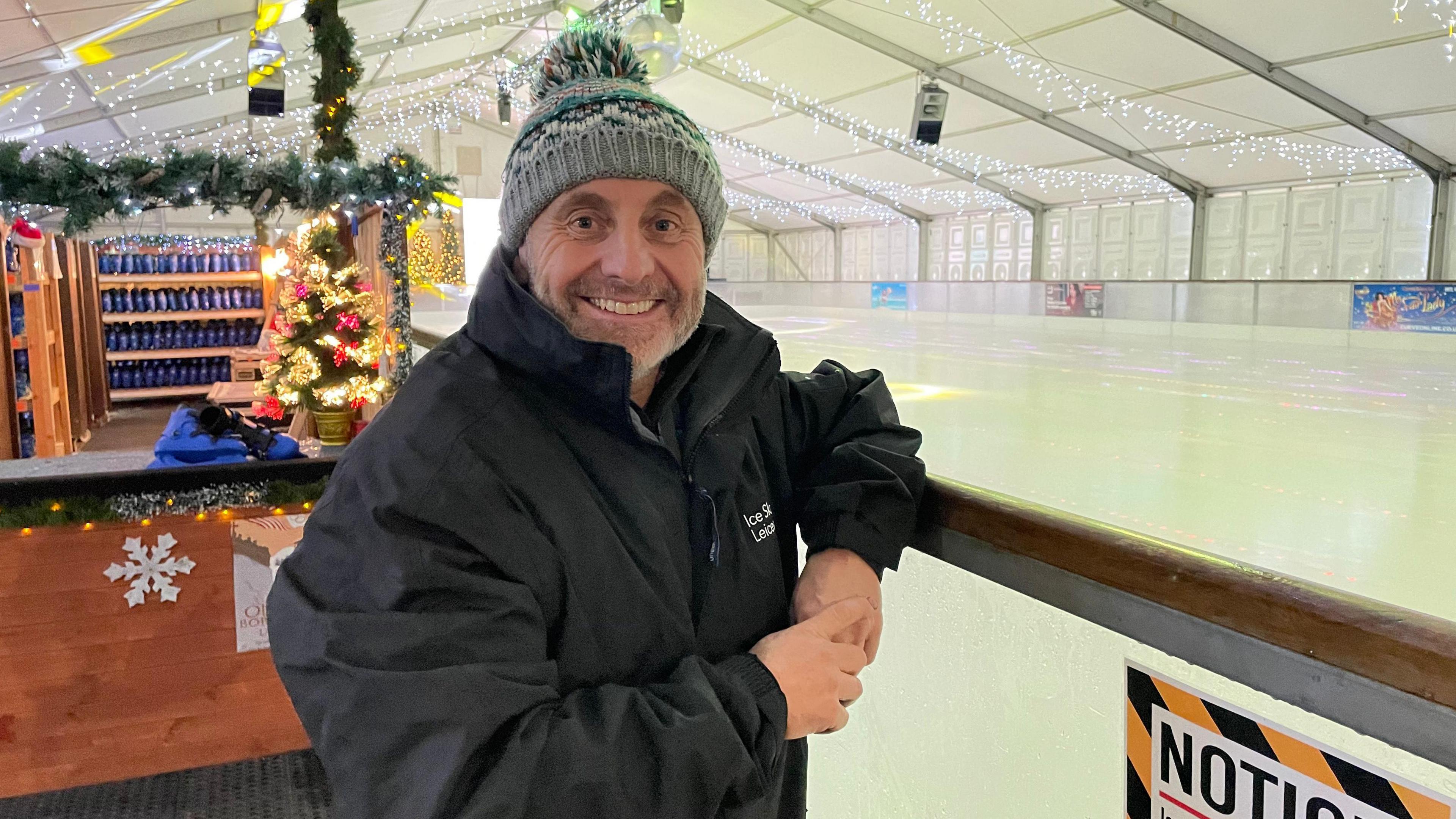 John Davies stood by an ice rink inside a marquee. He is wearing a light green bobble hat and black coat.