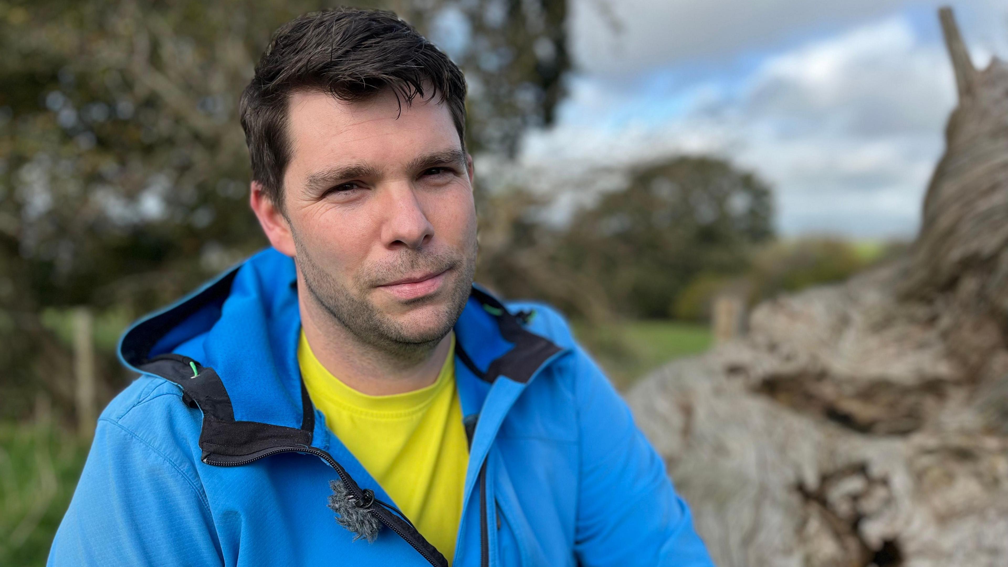 Michael Hall is sitting on a log in a field wearing a bright blue coat. He has dark brown hair and is staring right into the camera.