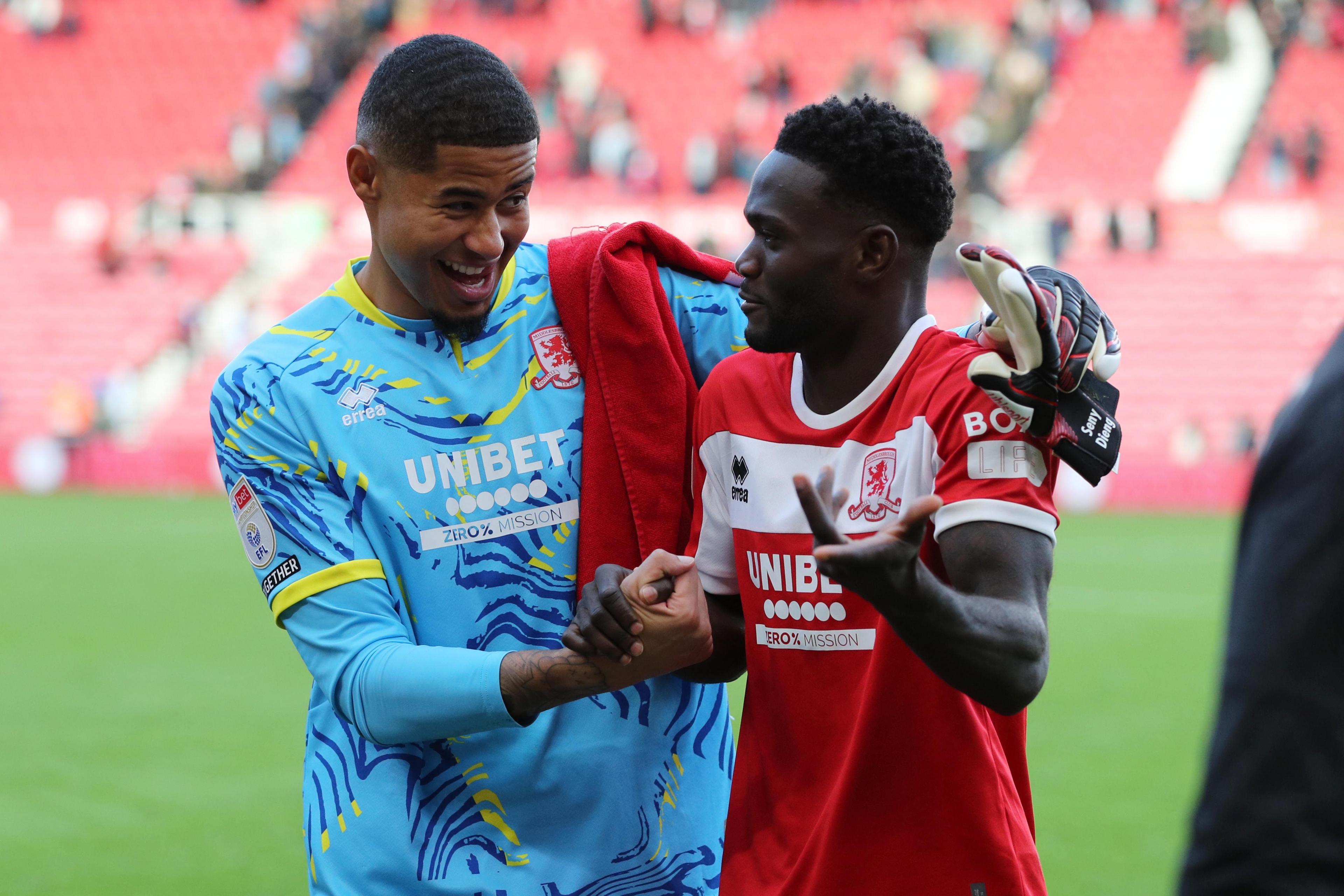 A goalkeeper in blue clasps hands with his teammate in a red shirt and smiles.