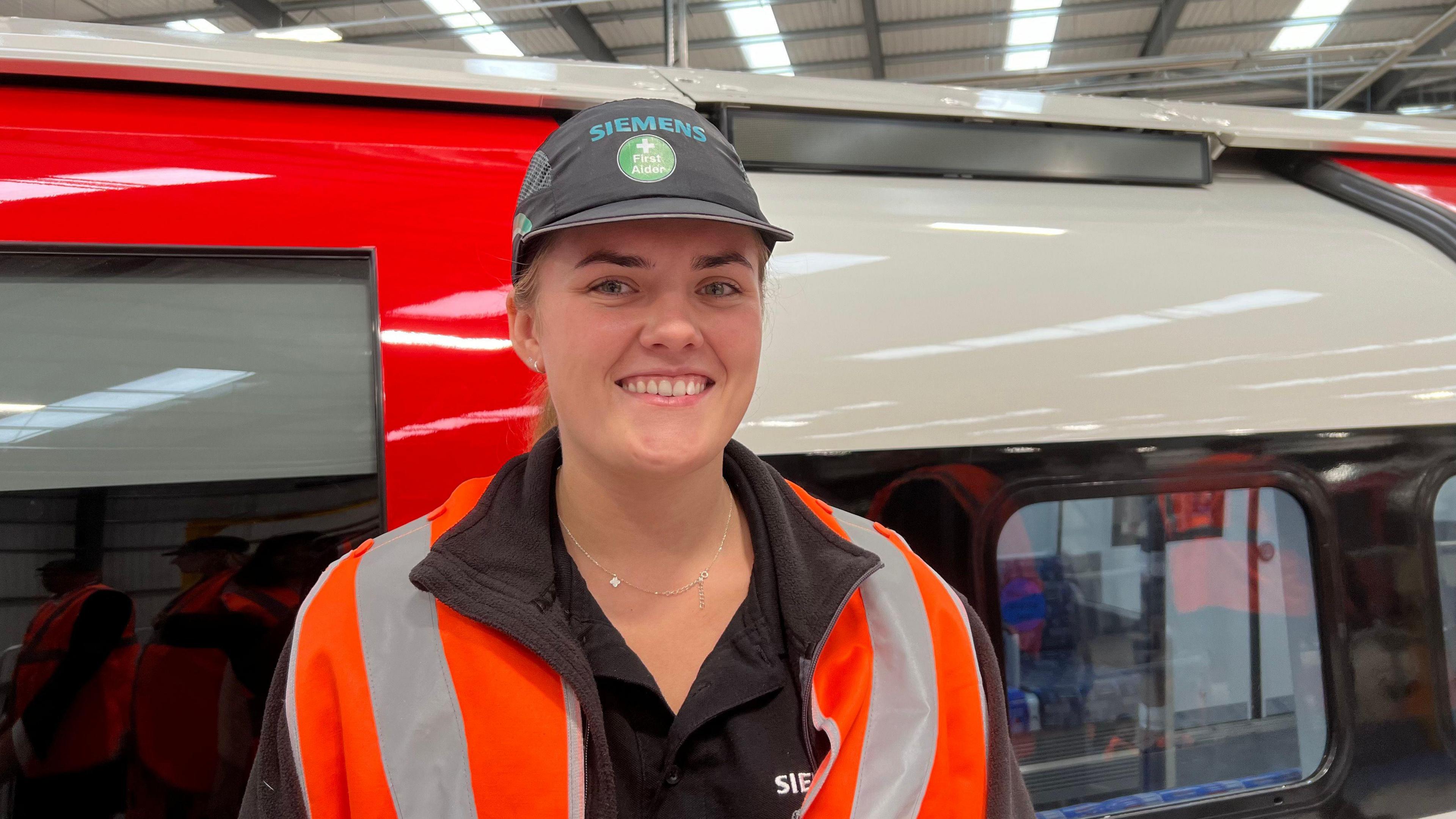 A woman is wearing a black Siemens' t-shirt with an orange hi-vis vest on and with a black Siemens' cap. She is smiling at the camera in front of a tube carriage that she is working on.