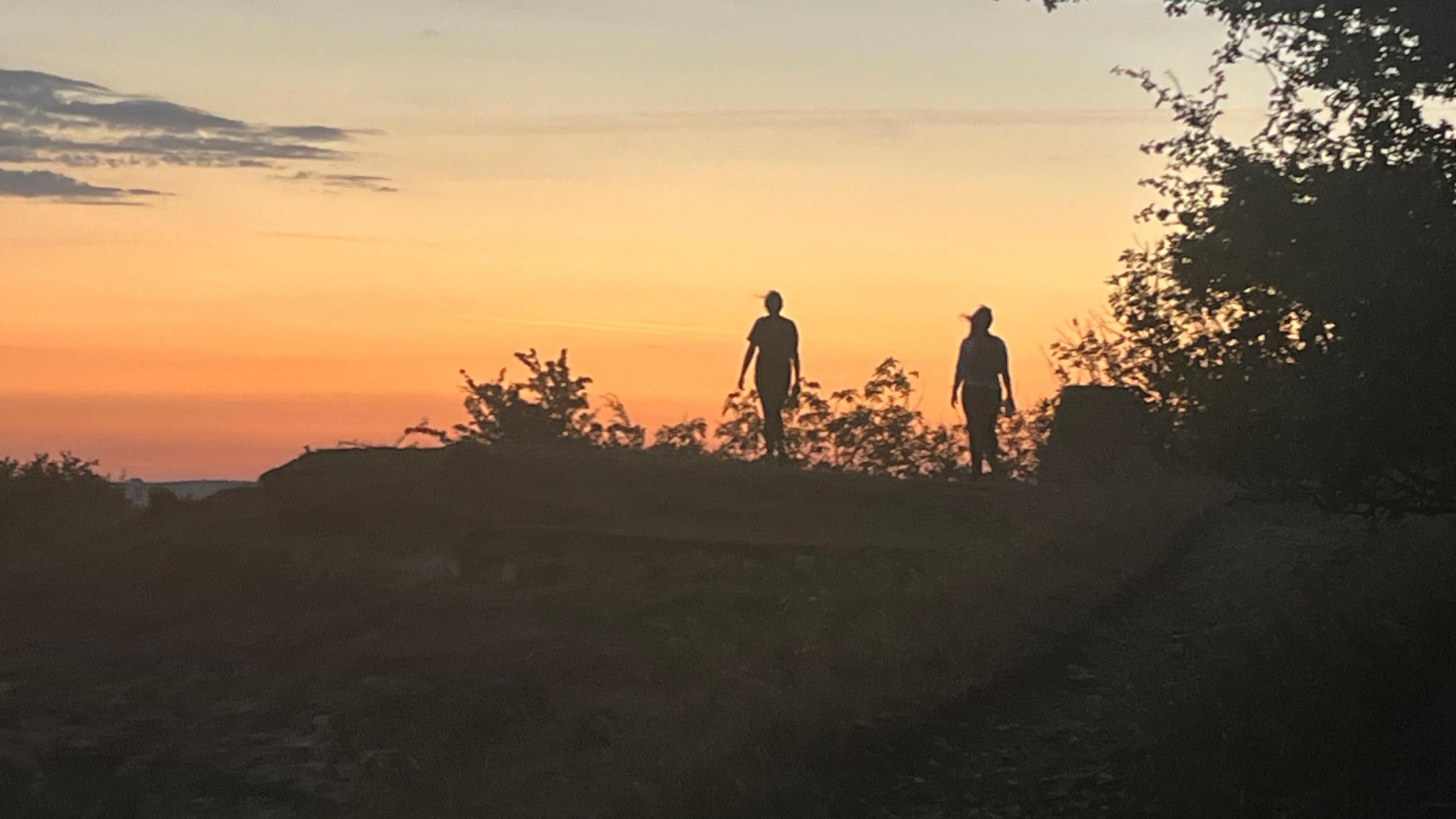 Two silhouetted people on the brow of a hill, walking during sunrise.