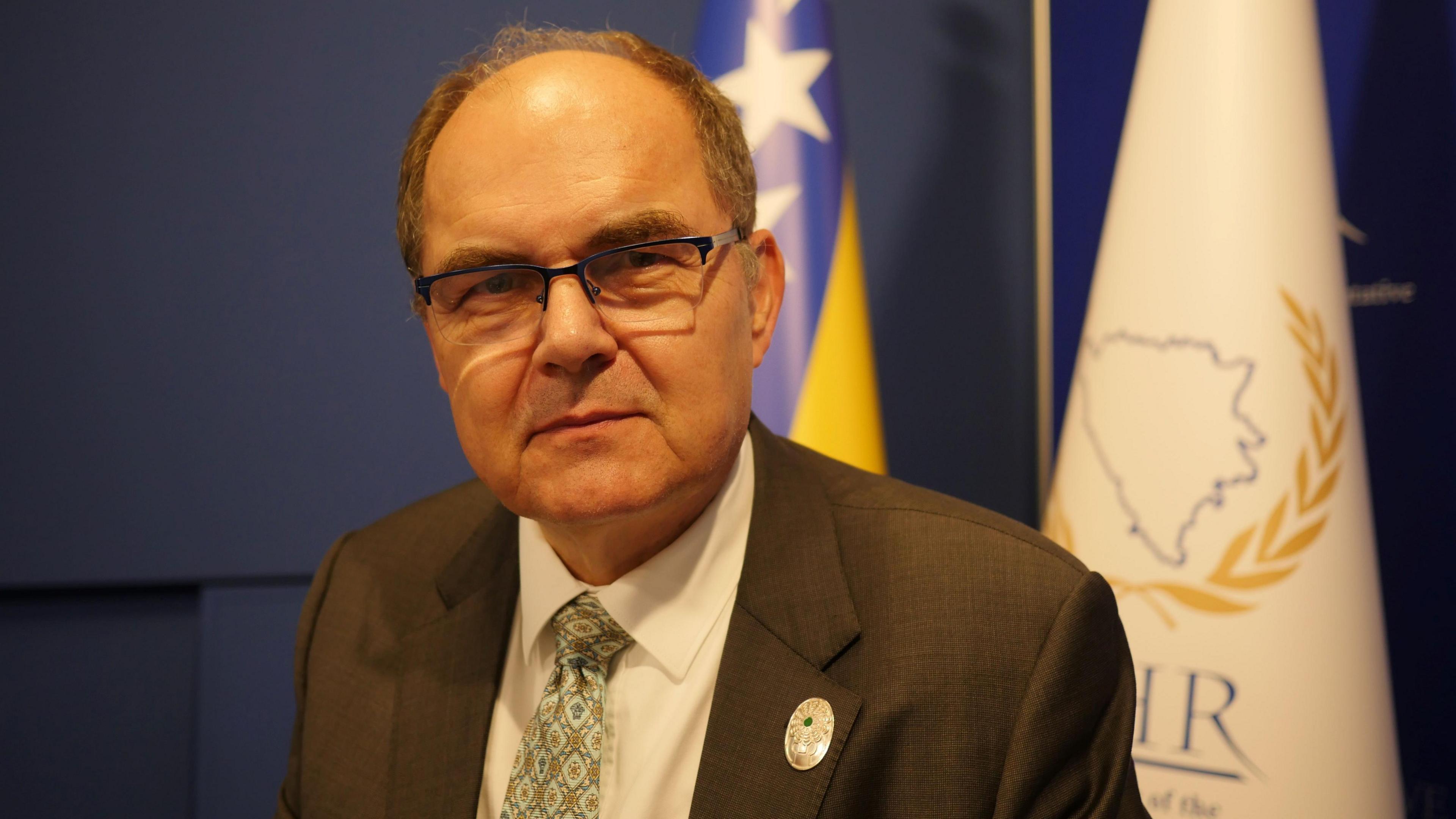 An older gentleman wearing glasses and a suit looks into the camera.