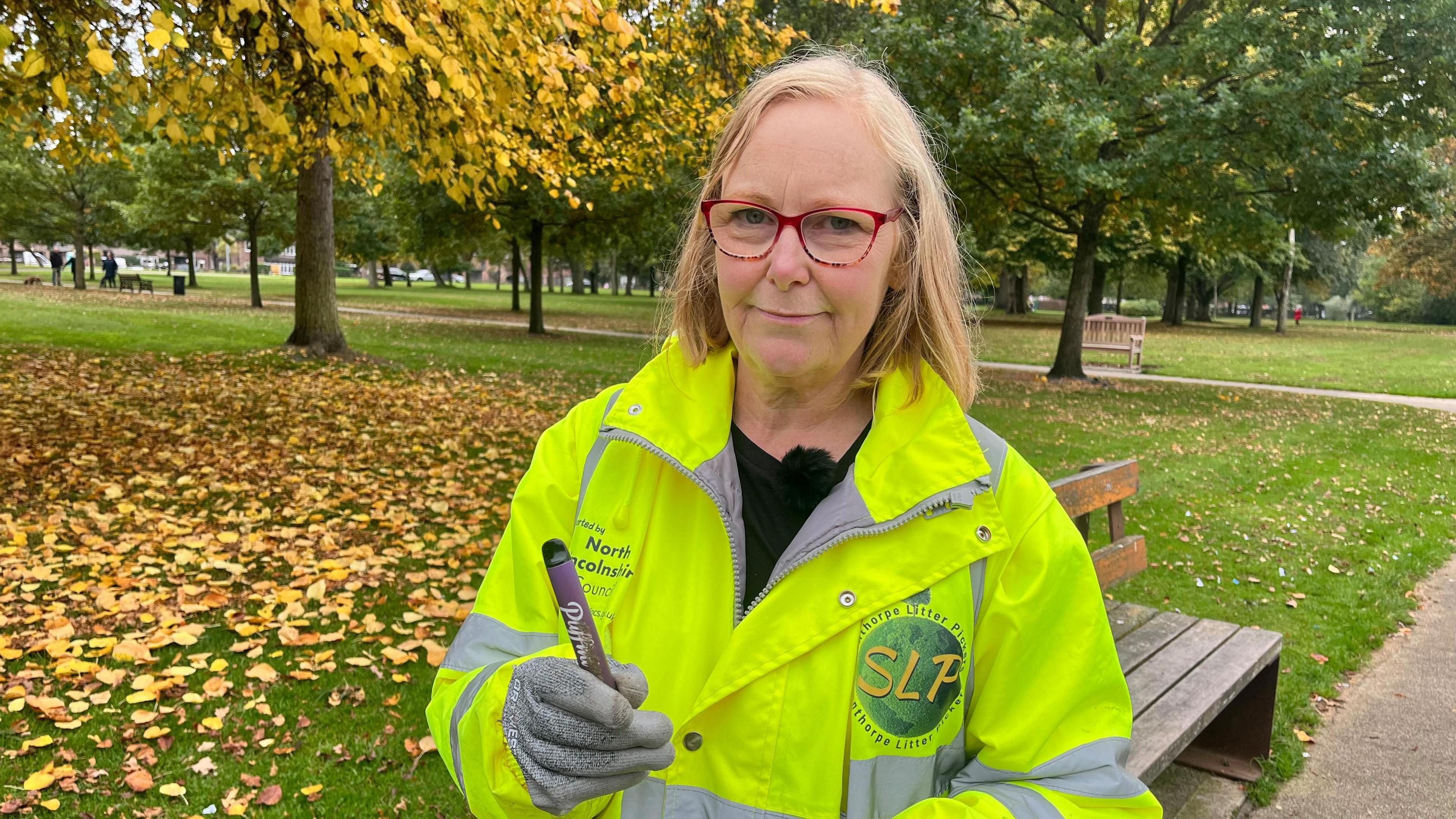 A woman with blond hair stands in Central Park in Scunthorpe with a bed of leaves on the grass behind her. She is wearing a bright yellow hi-vis jacket and holds a pen-sized disposable vape which she has just found in the park.