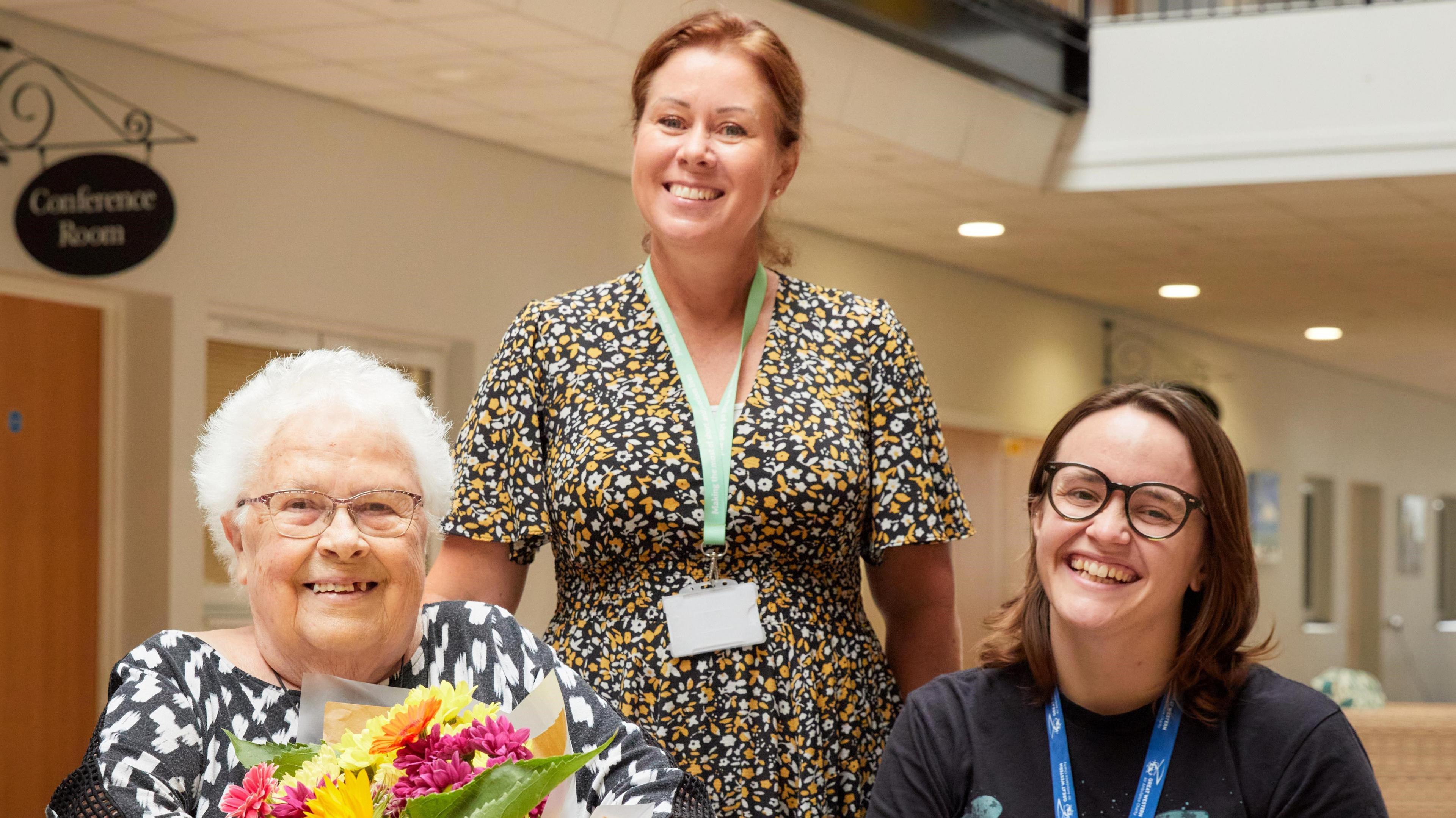 Former Gogglebox star Marine sits smiling along with two members of staff at the St Monica Trust home where she lives. She is wearing a black and white patterned dress and is holding a bunch of yellow, orange and pink flowers
