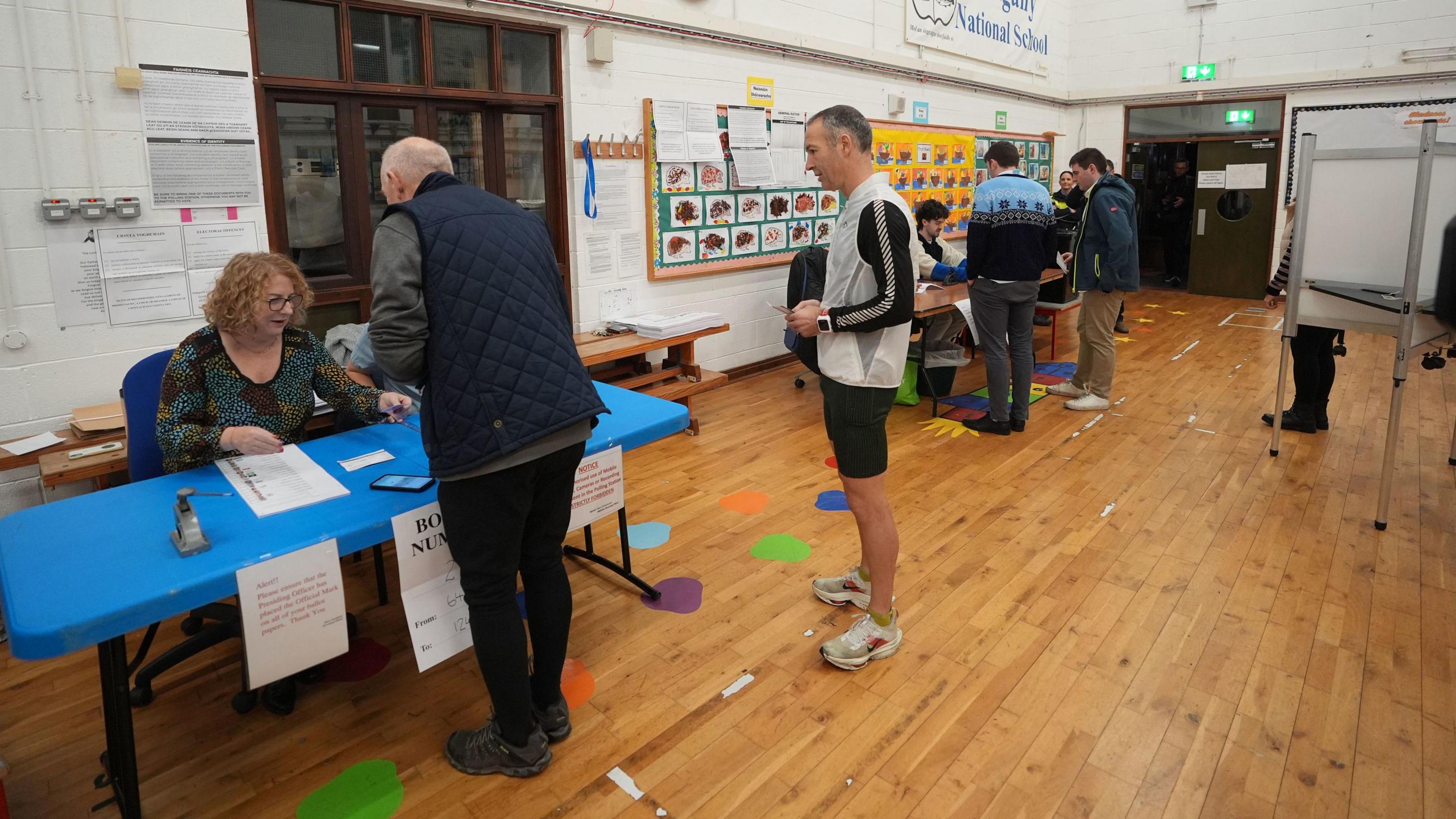 A woman sits at a blue desk in a school issuing voting cards. She has blonde hair and glasses. Two men line up at the desk. In the background there is a desk with two other men lined up to collect voting cards.