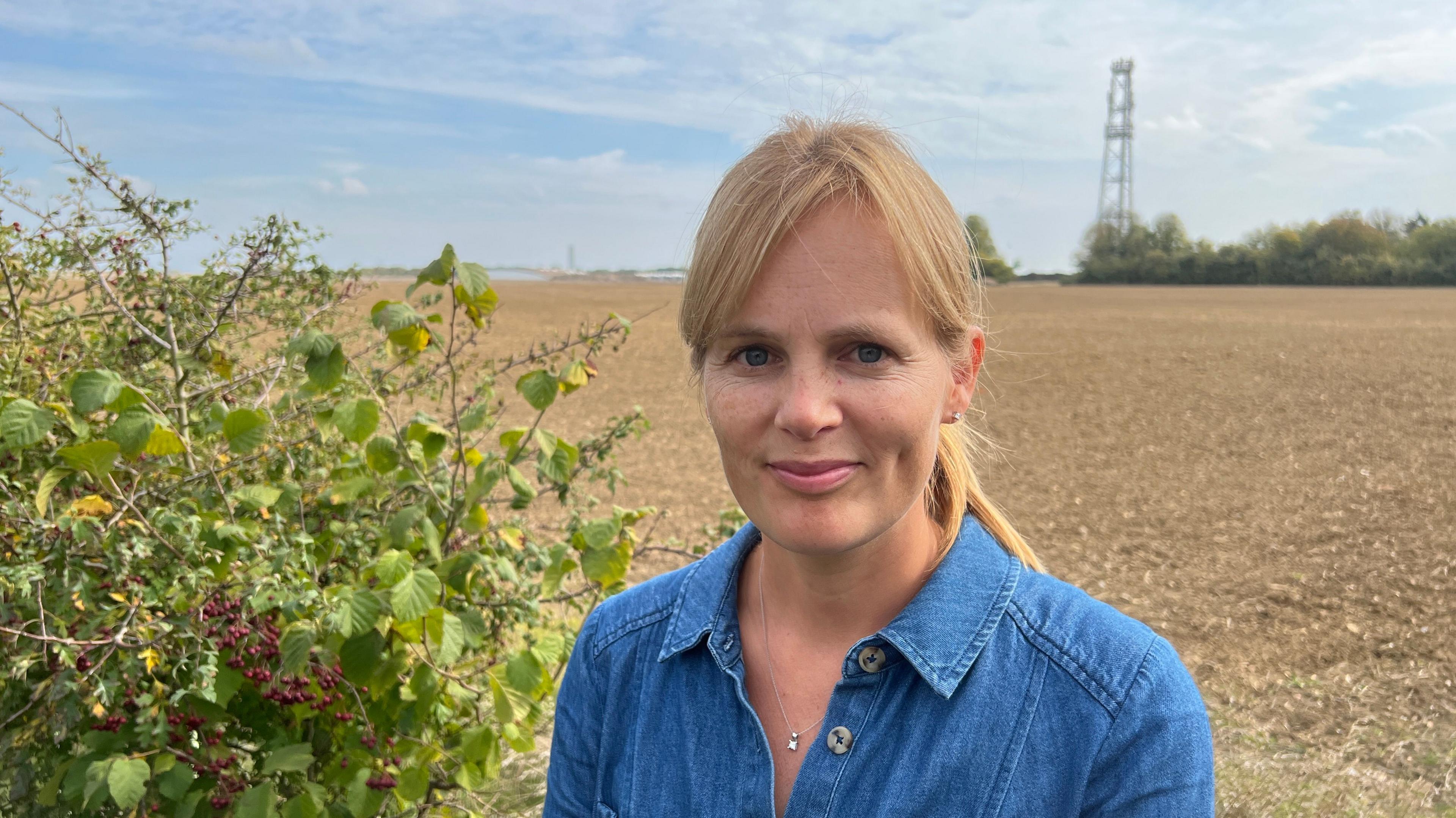 A fair-haired lady in her early forties smiling at the camera wearing a blue denim top.  She is standing in front of a field and berry bush.  In the distance a factory plant can be seen faintly.