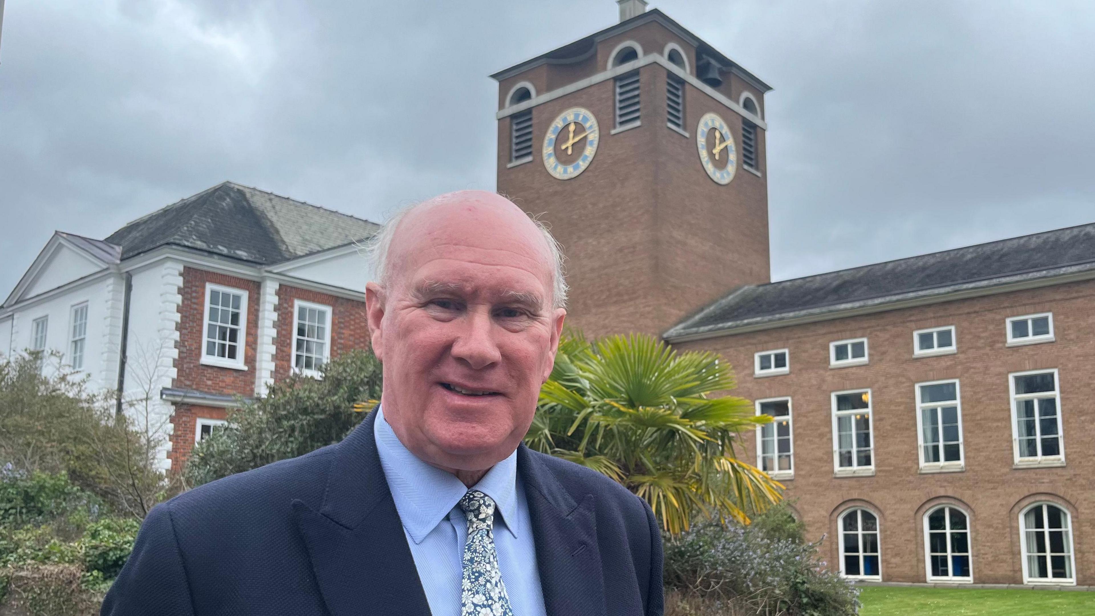 The Conservative leader of Devon County Council, James McInnes, wearing a blue shirt and jacket and a floral tie, standing in front of the clock tower at County Hall.