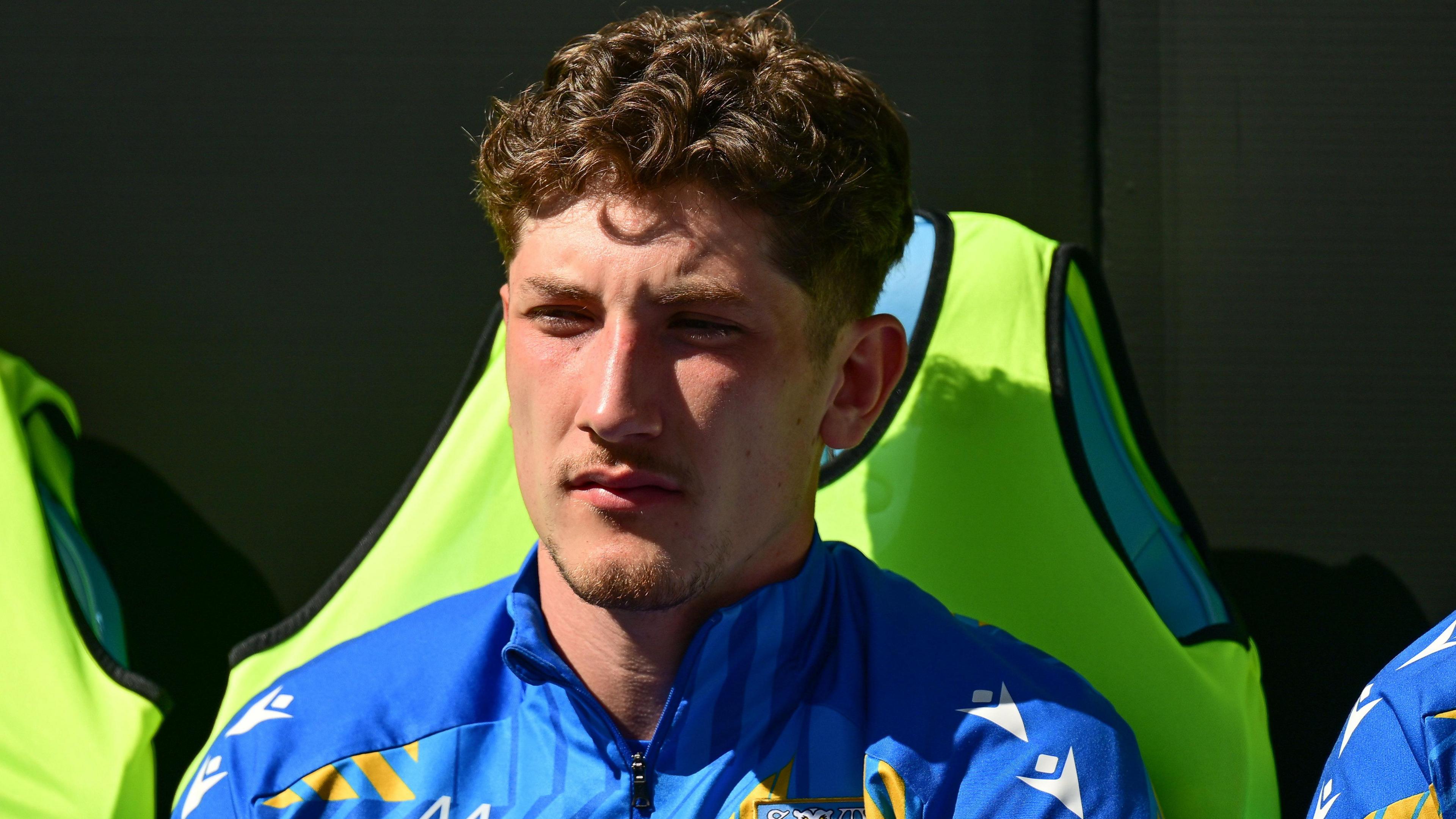 Mackenzie Maltby, in a blue tracksuit top, sitting on a green pitchside seat.