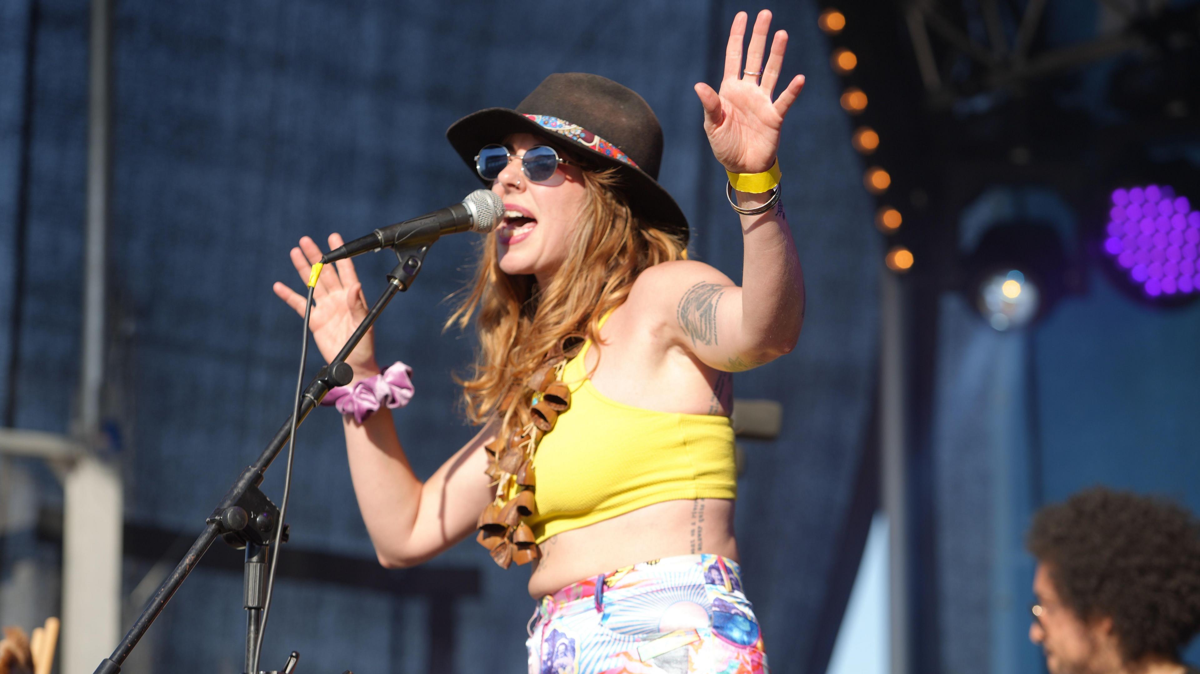 Woman performing on stage. Her arms are in the air and she is singing into a mic. She is wearing a hat and sunglasses. 