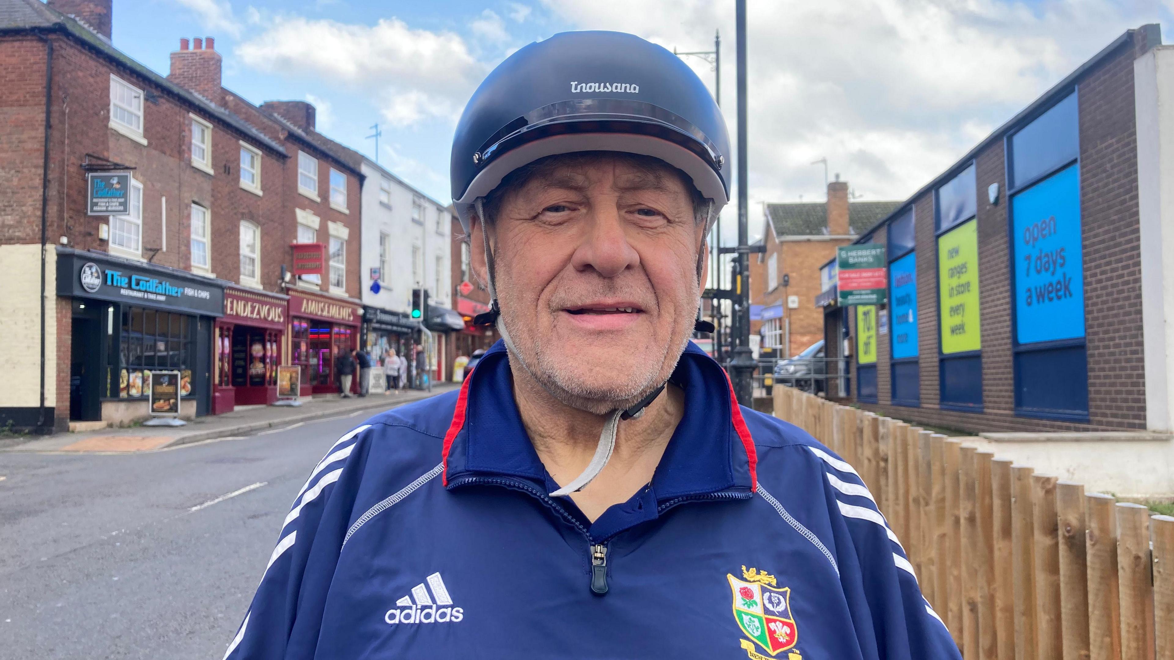 A man stood down a road, wearing a blue sports jacket and a cycle hat.