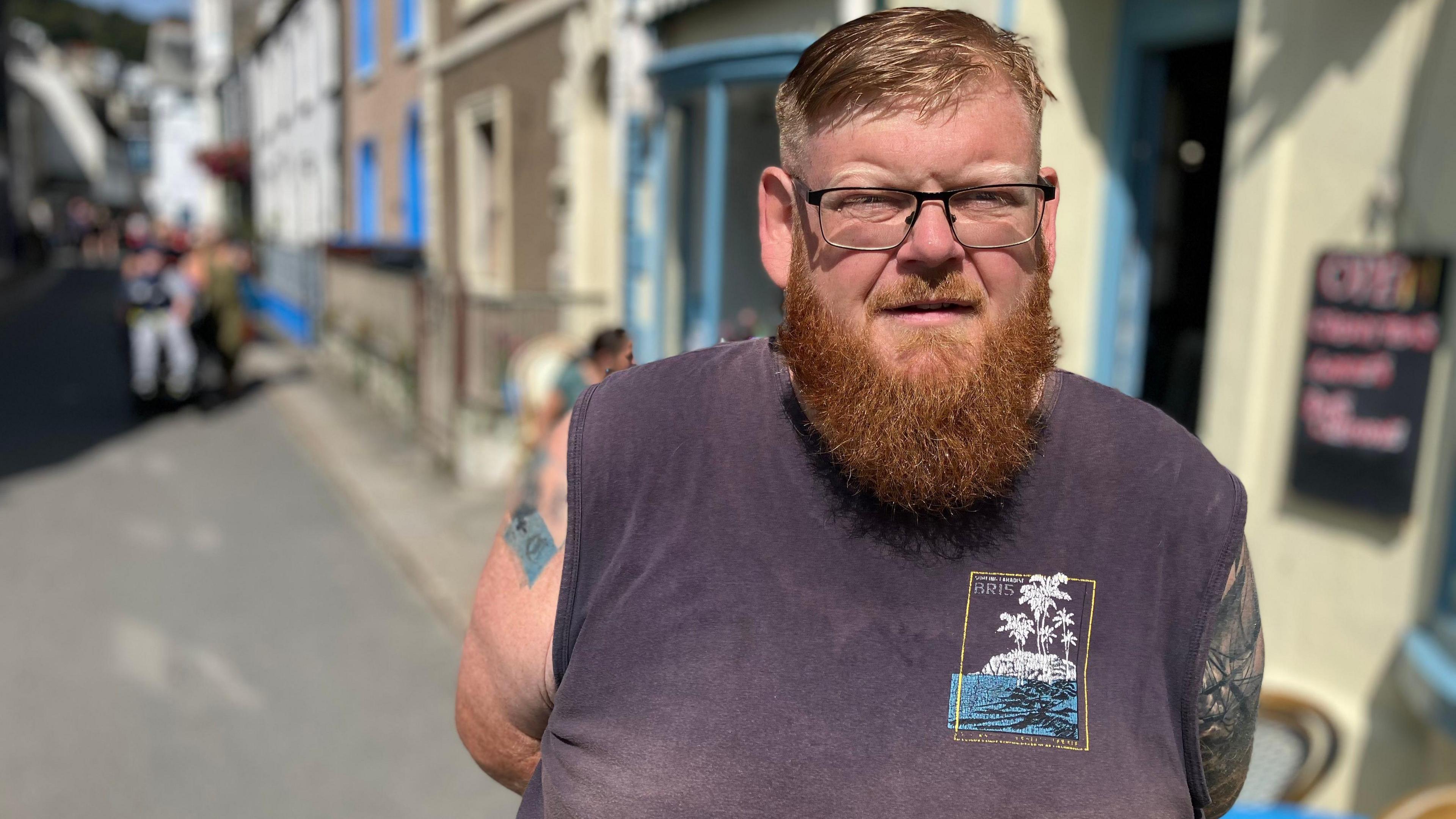 A man stands in a the street in a Cornish town, it is sunny and he is wearing a vest top.