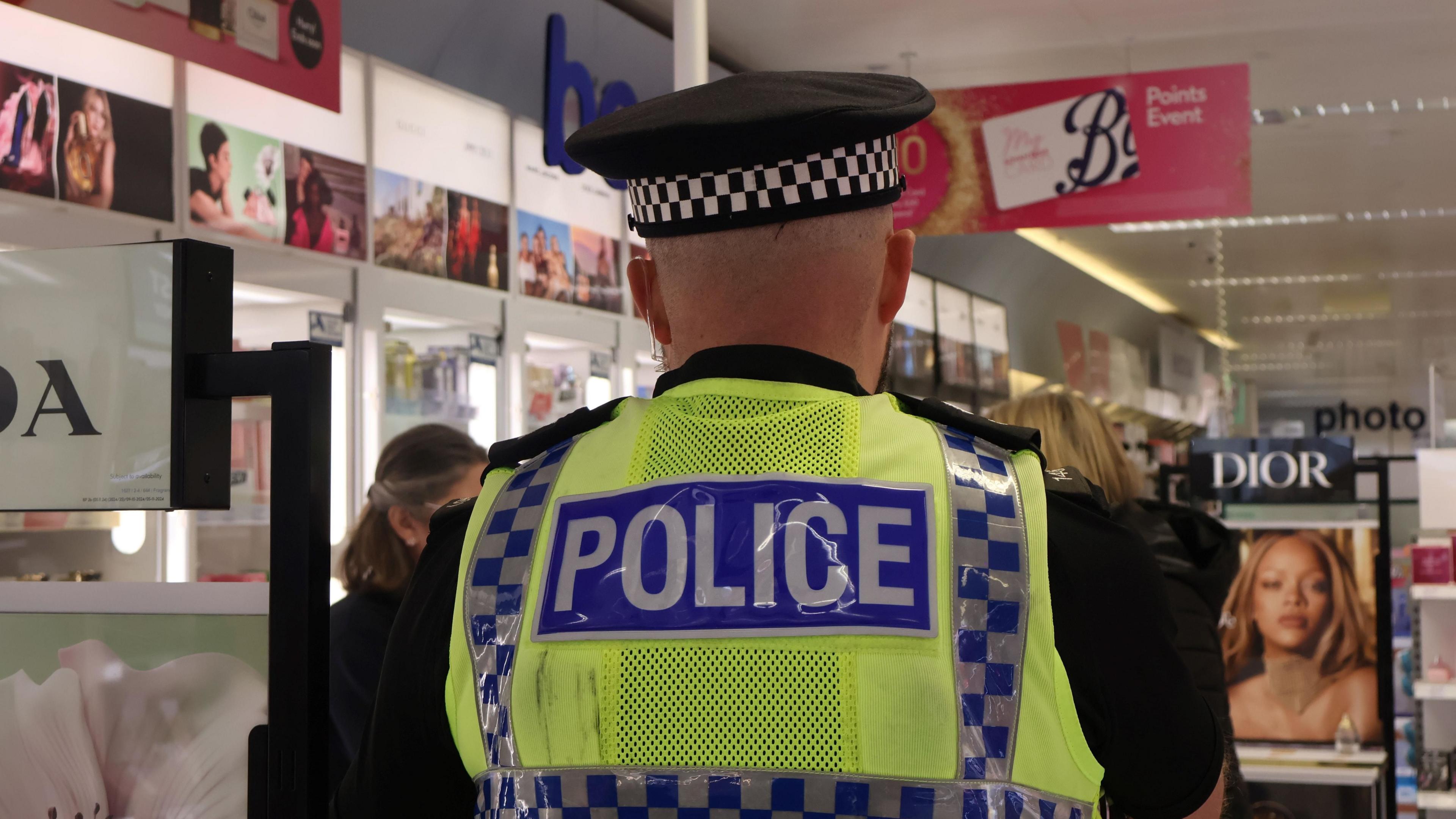 A police officer facing away from the camera in a shop. He is wearing a black police hat with black and white squares on it. A black short sleeve shirt and a high visibility police vest. Behind him are advertising hoardings and shop staff. 