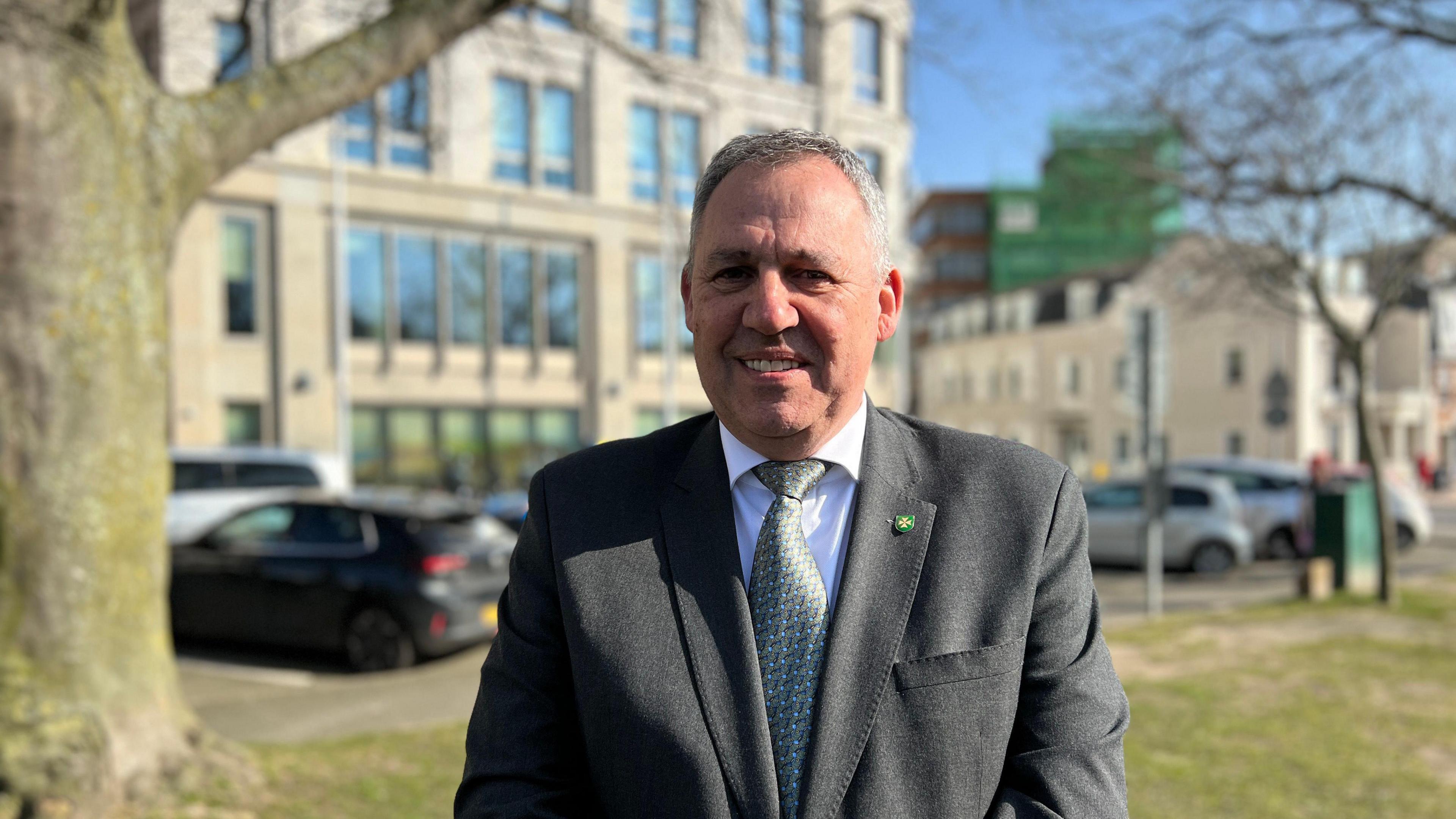 Andy Jehan seen wearing a grey suit with a white shirt and green patterned tie. He has short grey hair and is stood outside near a street with cars and a large tree to his right.