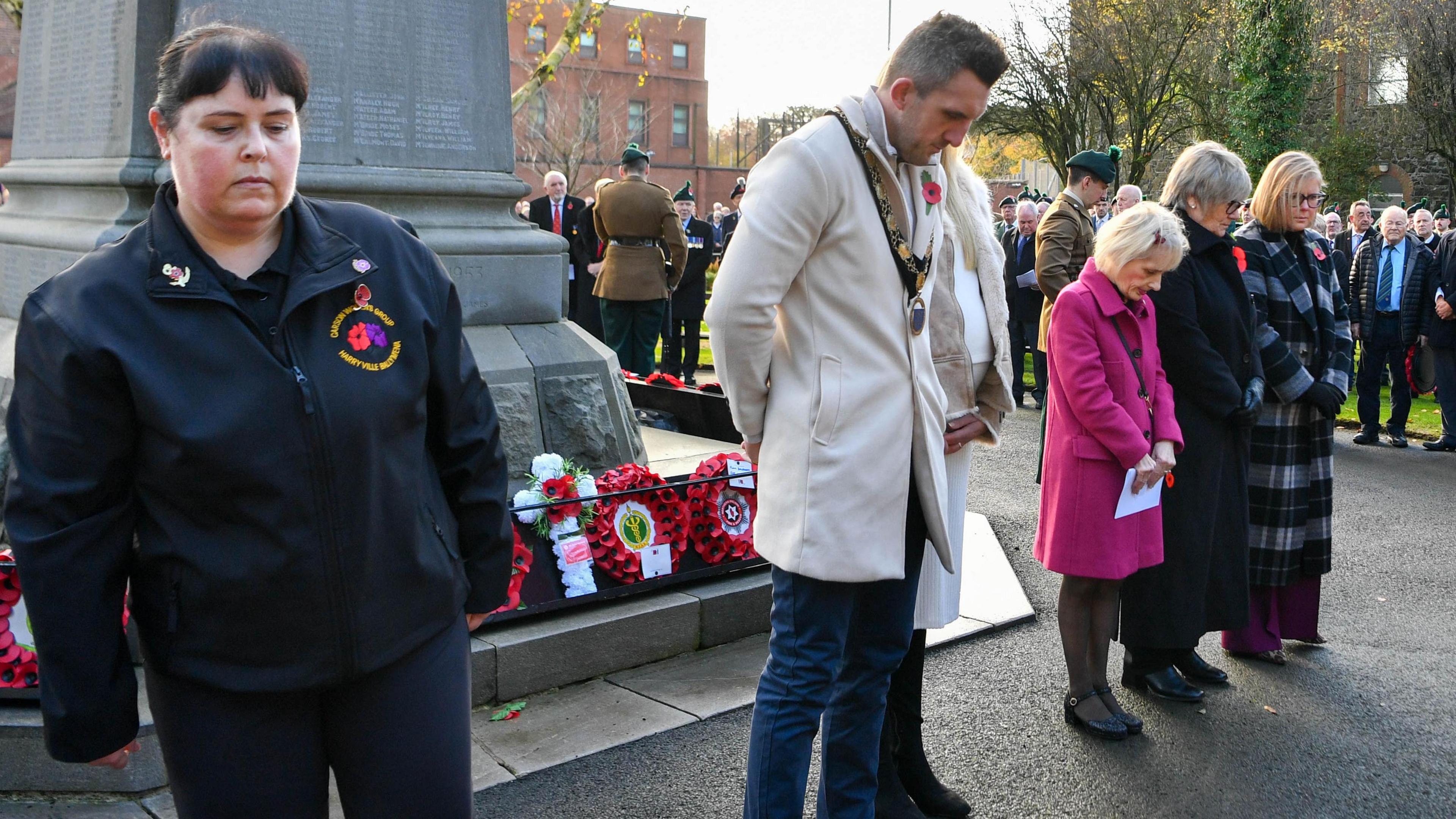 People are looking at the ground as they hold two minutes of silence. Some are wearing poppies. Wreaths are sitting around a memorial. 