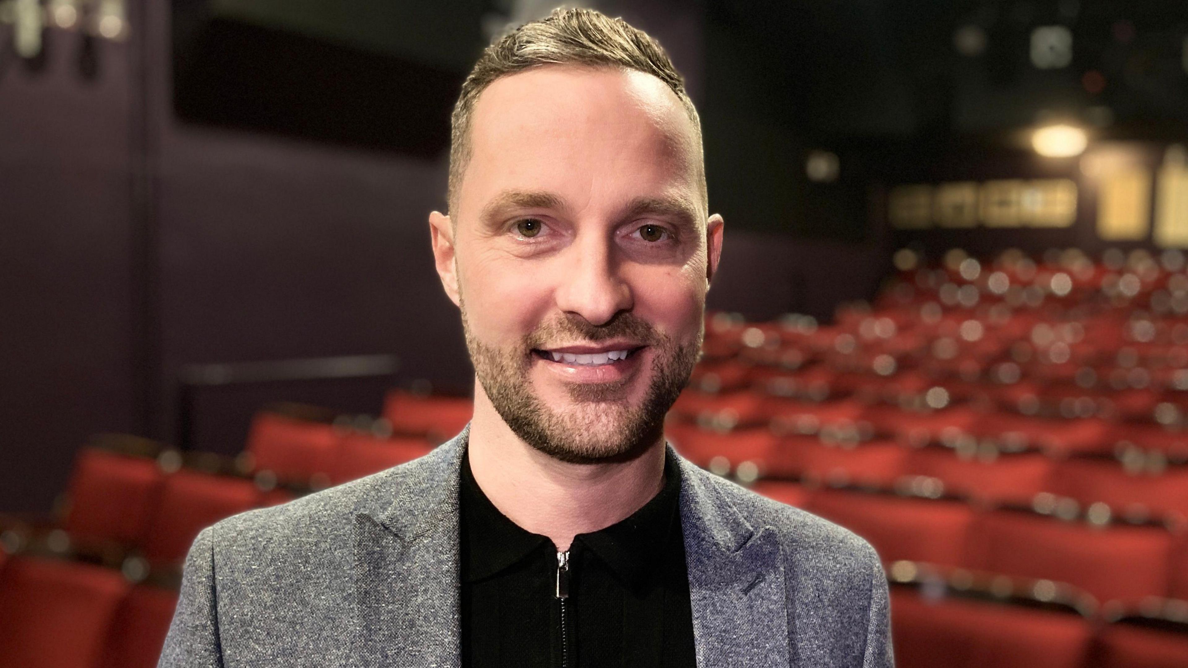 Man with stubble stood in front of red seats in cinema