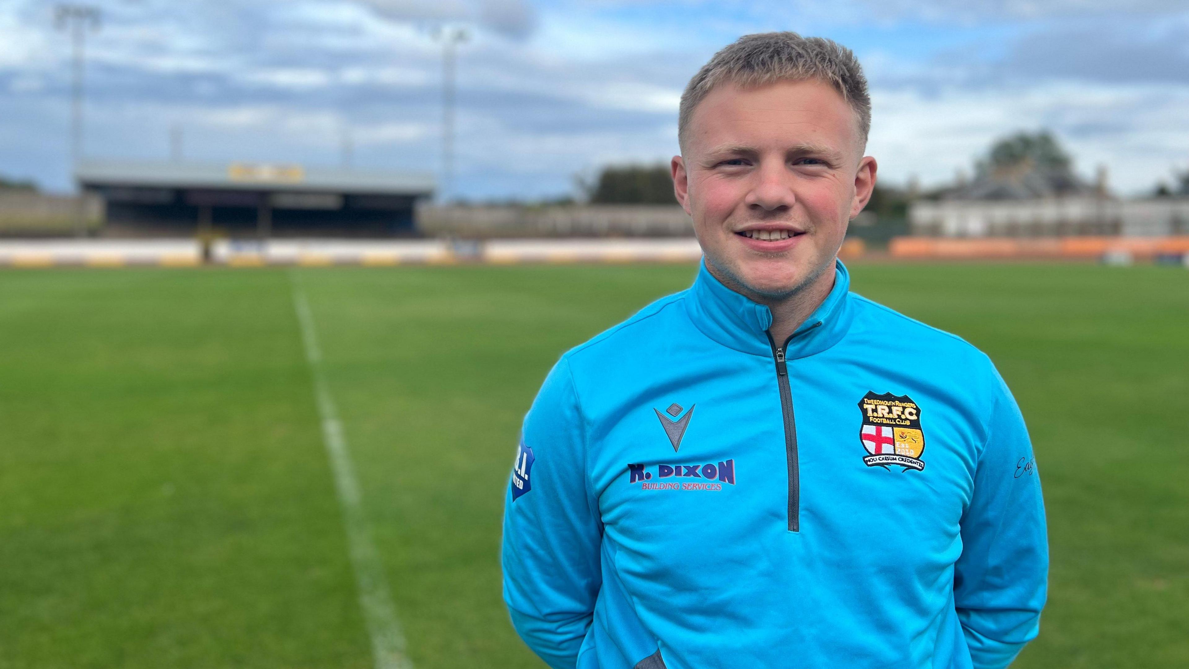 A man with light brown hair and wearing a blue sports top stands in front of a football pitch