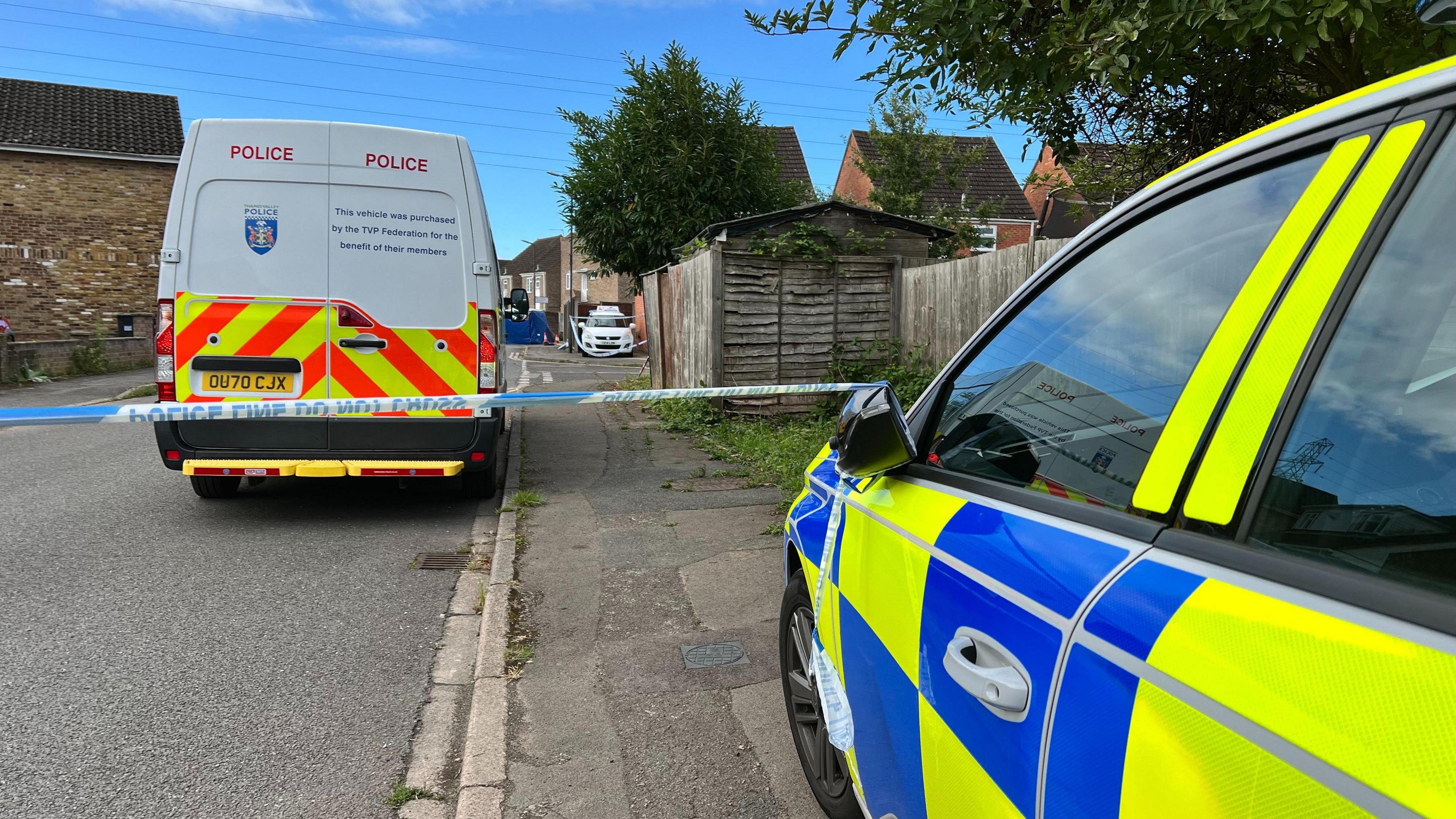 The side of a police car stopped just before a strip of blue and white police tape stretched across a residential street, with a police van behind the cordon.